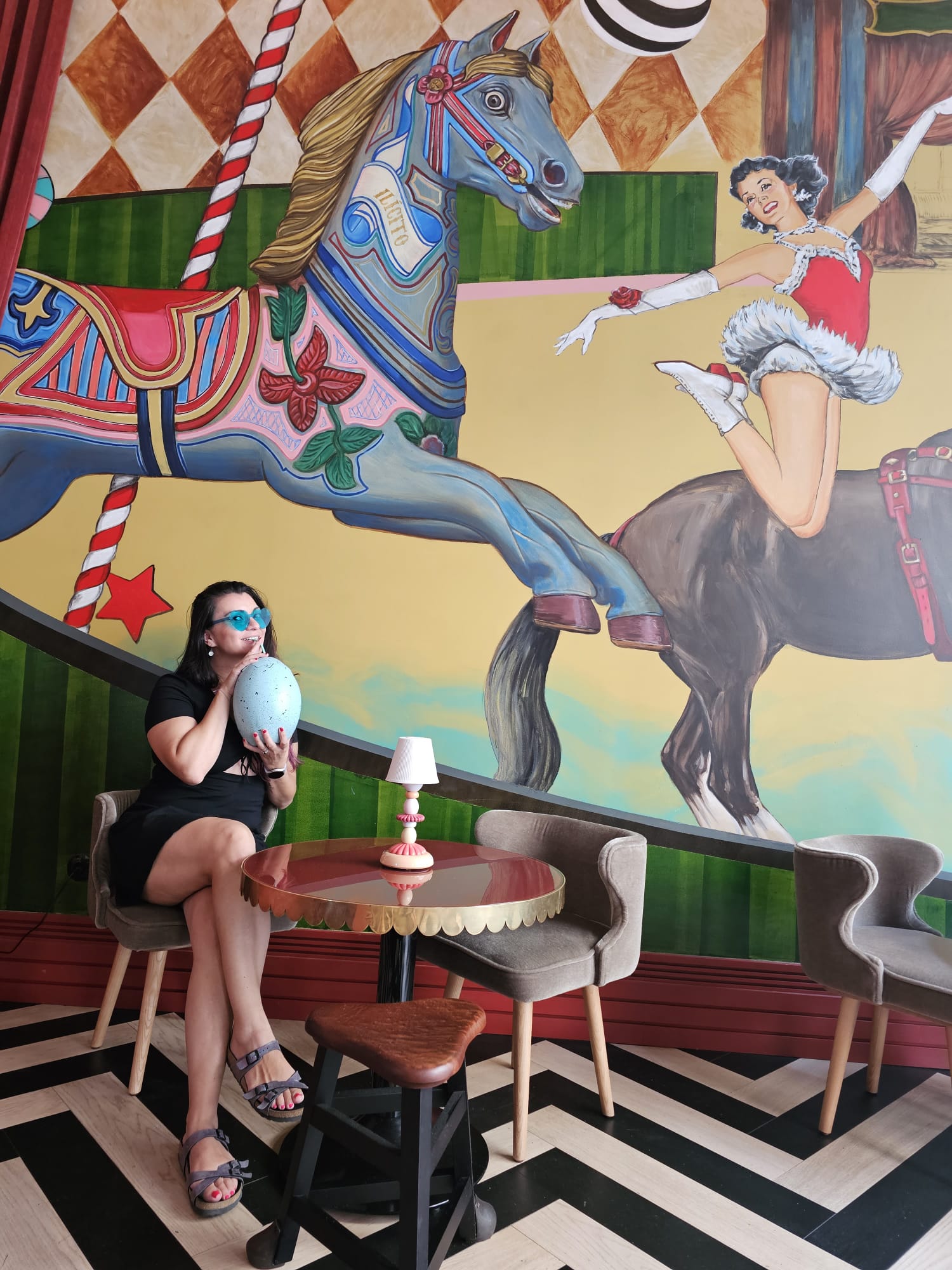 A woman in a black dress sits at a small round table, holding a decorative egg, in front of a vibrant mural featuring a carousel horse and an acrobat. The setting includes modern chairs, a small lamp on the table, and a bold zigzag floor pattern.