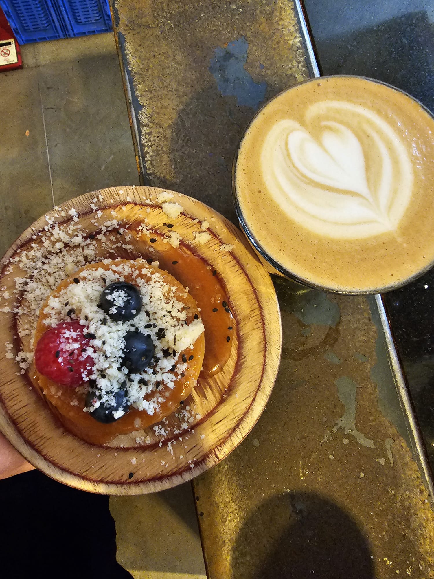 A latte with heart-shaped foam art sits next to a pastry topped with fresh raspberries, blueberries, and grated cheese on a wooden plate.