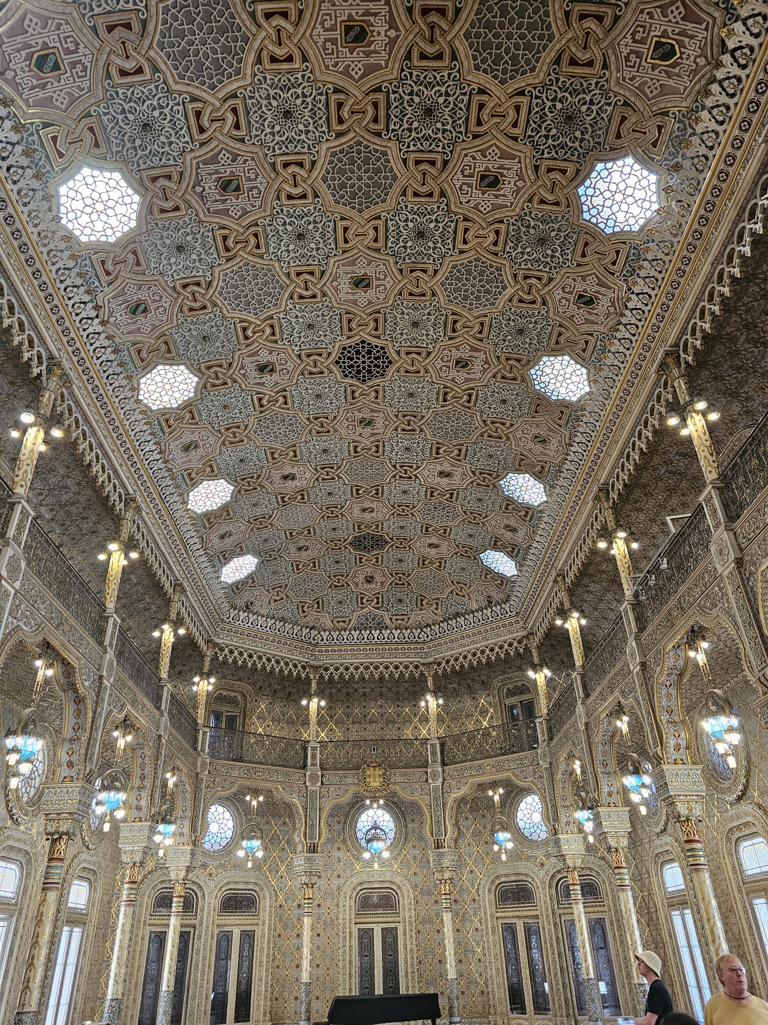 The image showcases a grand interior with an intricately patterned ceiling featuring geometric designs and hexagonal skylights. The walls are adorned with ornate arches and columns, and elegant chandeliers hang throughout the space. The room exudes a sense of opulence and architectural beauty.