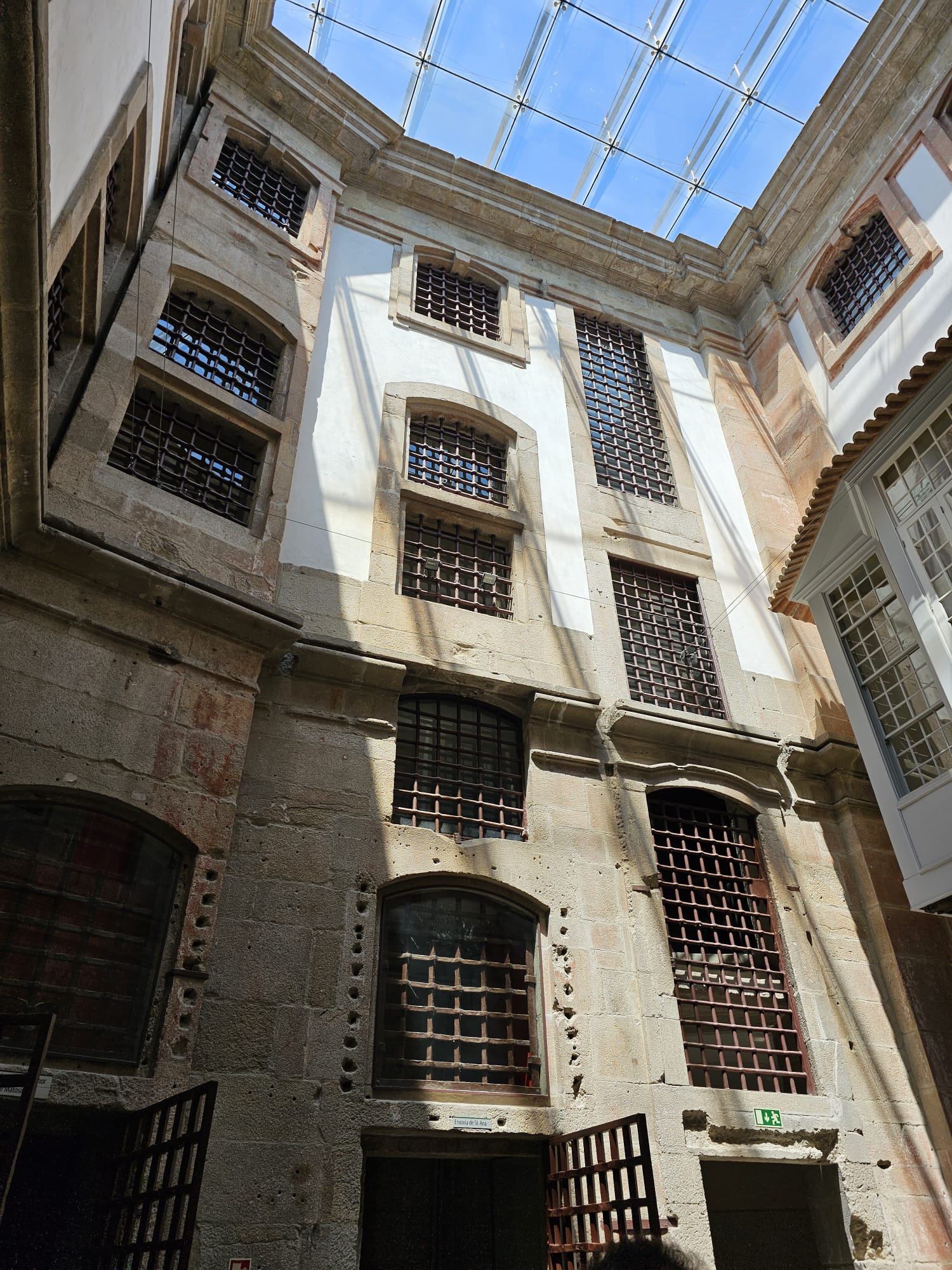 The image shows the interior courtyard of an old stone building with multiple barred windows and a glass ceiling. Sunlight streams through the transparent roof, casting shadows on the walls. The architecture features a mix of stone and white plaster, with an open doorway at the bottom.