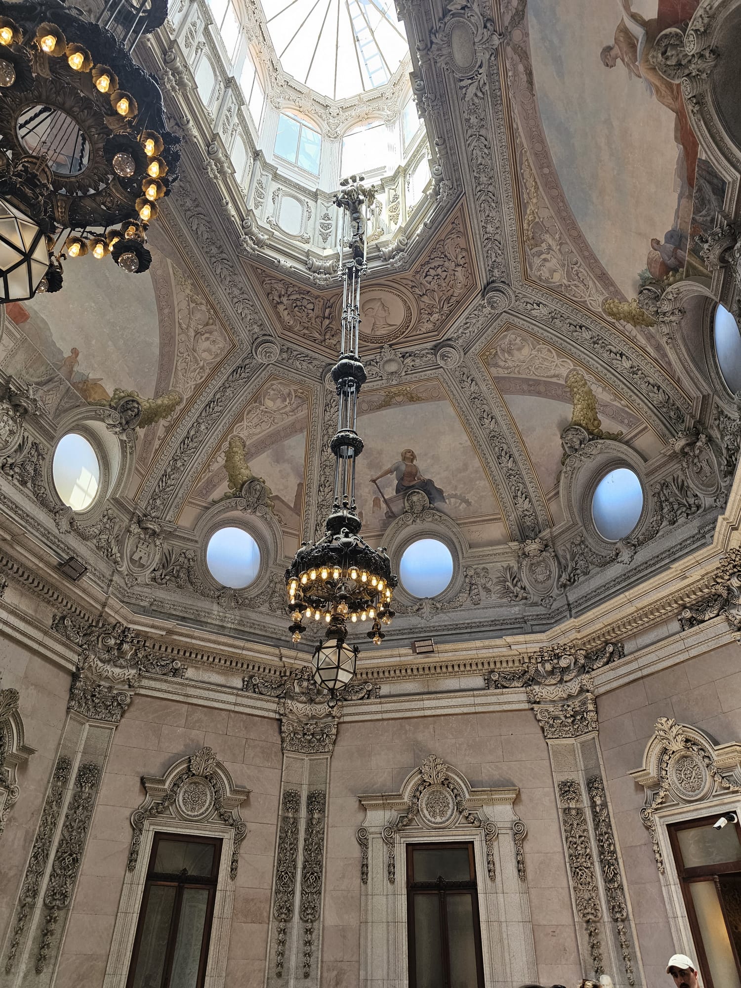 An ornate domed ceiling features intricate architectural details and classical frescoes, with a grand chandelier hanging from the center. The ceiling is adorned with decorative moldings and circular windows, allowing natural light to illuminate the elegant space below.