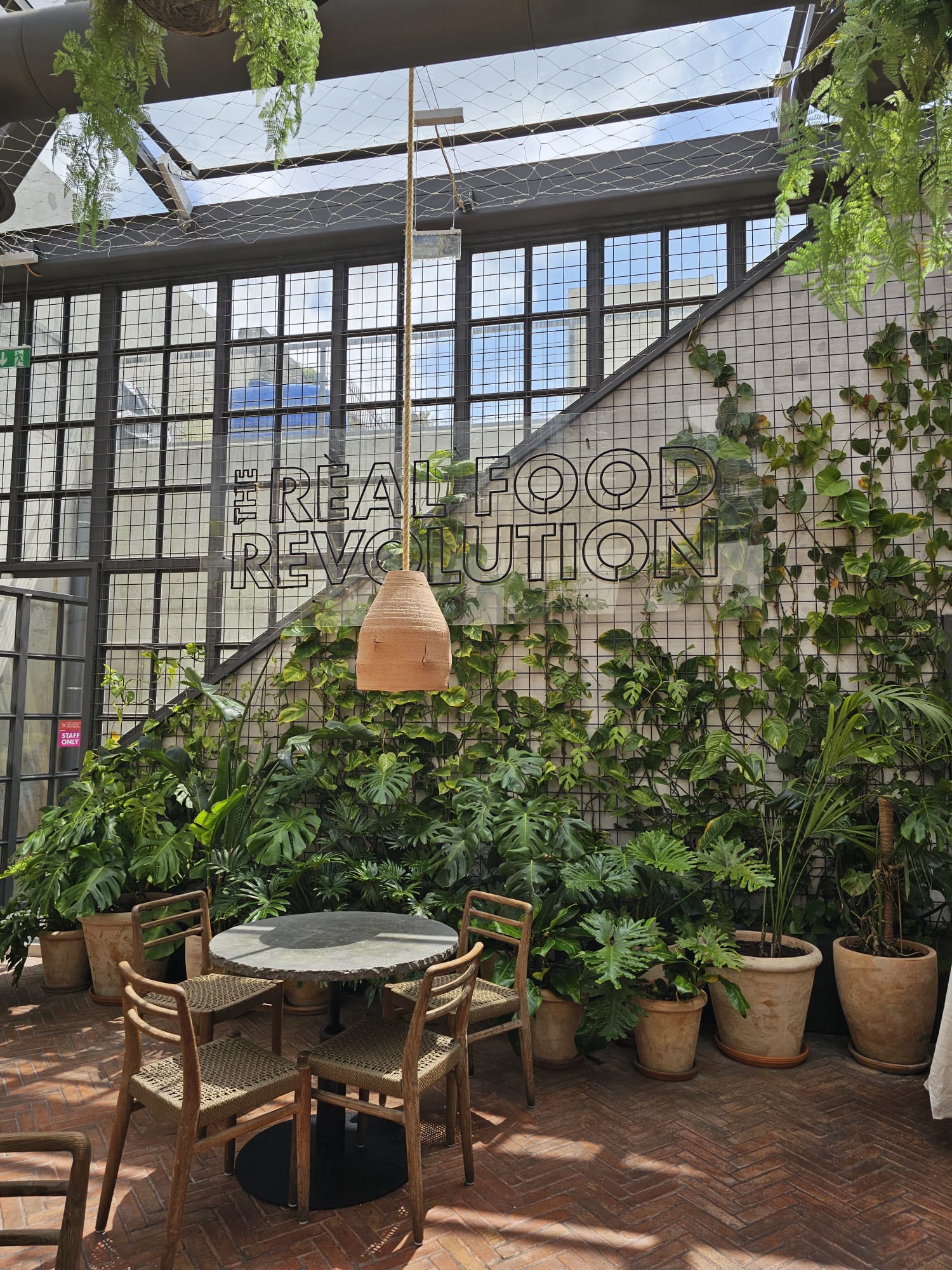 A cozy indoor patio features a round table with four wooden chairs, surrounded by lush potted plants. The wall is adorned with a vertical garden and the phrase "The Real Food Revolution" in large letters. Sunlight filters through a glass ceiling, highlighting the earthy brick floor.
