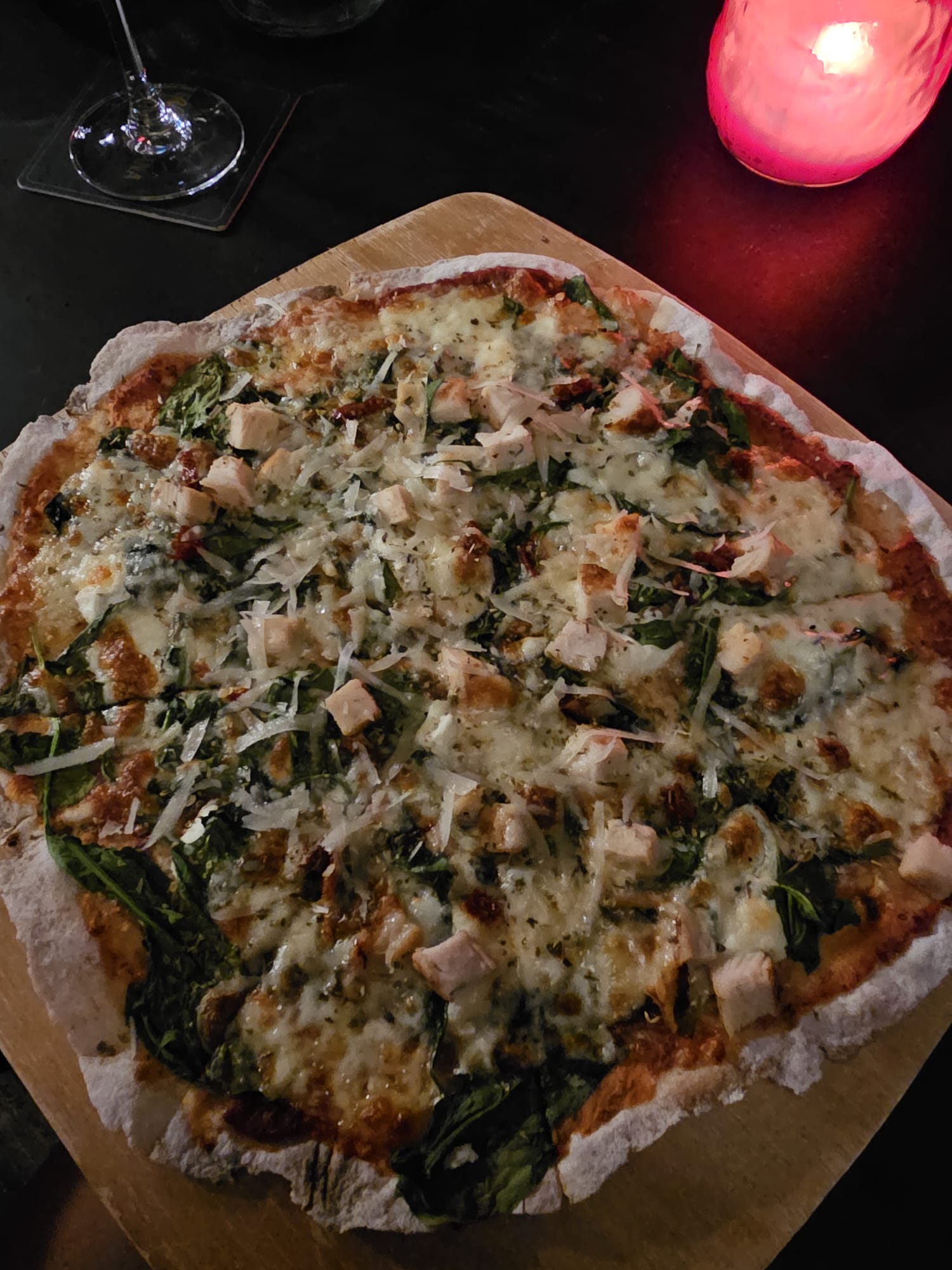 A freshly baked pizza topped with melted cheese, spinach, and chunks of chicken sits on a wooden board. A glowing red candle and a wine glass are visible in the dimly lit background, creating a cozy atmosphere.