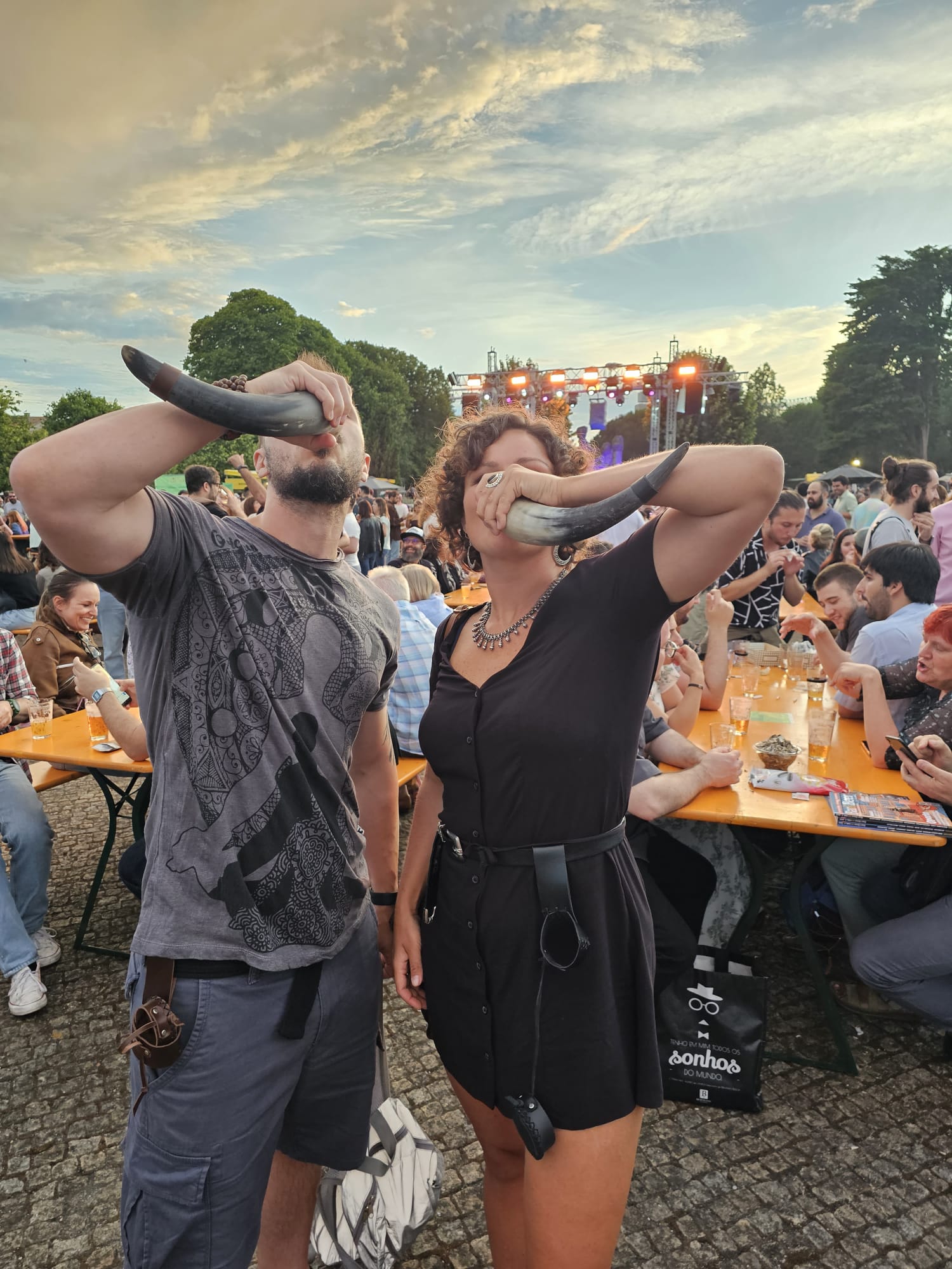 Two people are drinking from large horns at an outdoor event, surrounded by a lively crowd seated at picnic tables. The sky is partly cloudy, and a stage with lights is visible in the background.