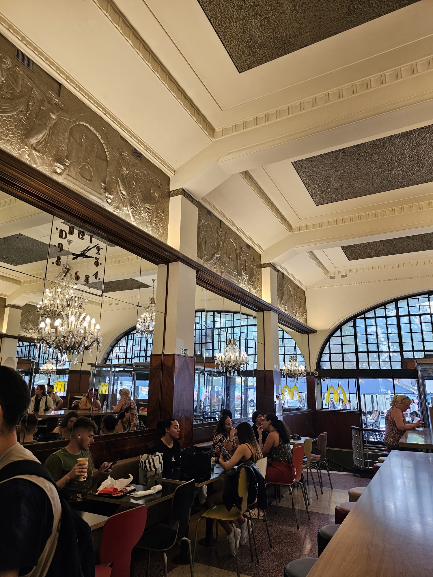 A bustling fast-food restaurant with ornate decor, featuring large chandeliers and decorative wall reliefs. Patrons sit at tables, eating and chatting, while others stand near the counter. Large windows let in natural light, and the iconic golden arches logo is visible outside.