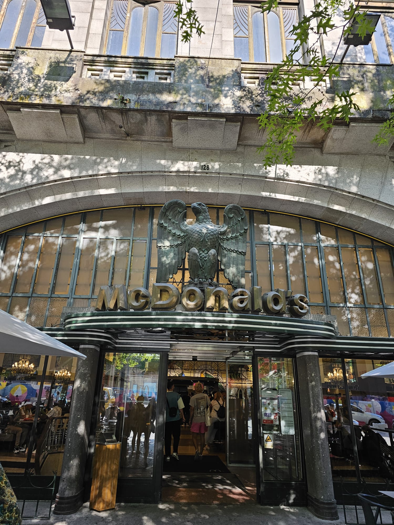 The entrance to a McDonald's restaurant features an ornate facade with large glass windows and a prominent eagle sculpture above the logo. People are entering the building, and outdoor seating is visible under umbrellas. Shadows from nearby trees dapple the scene.