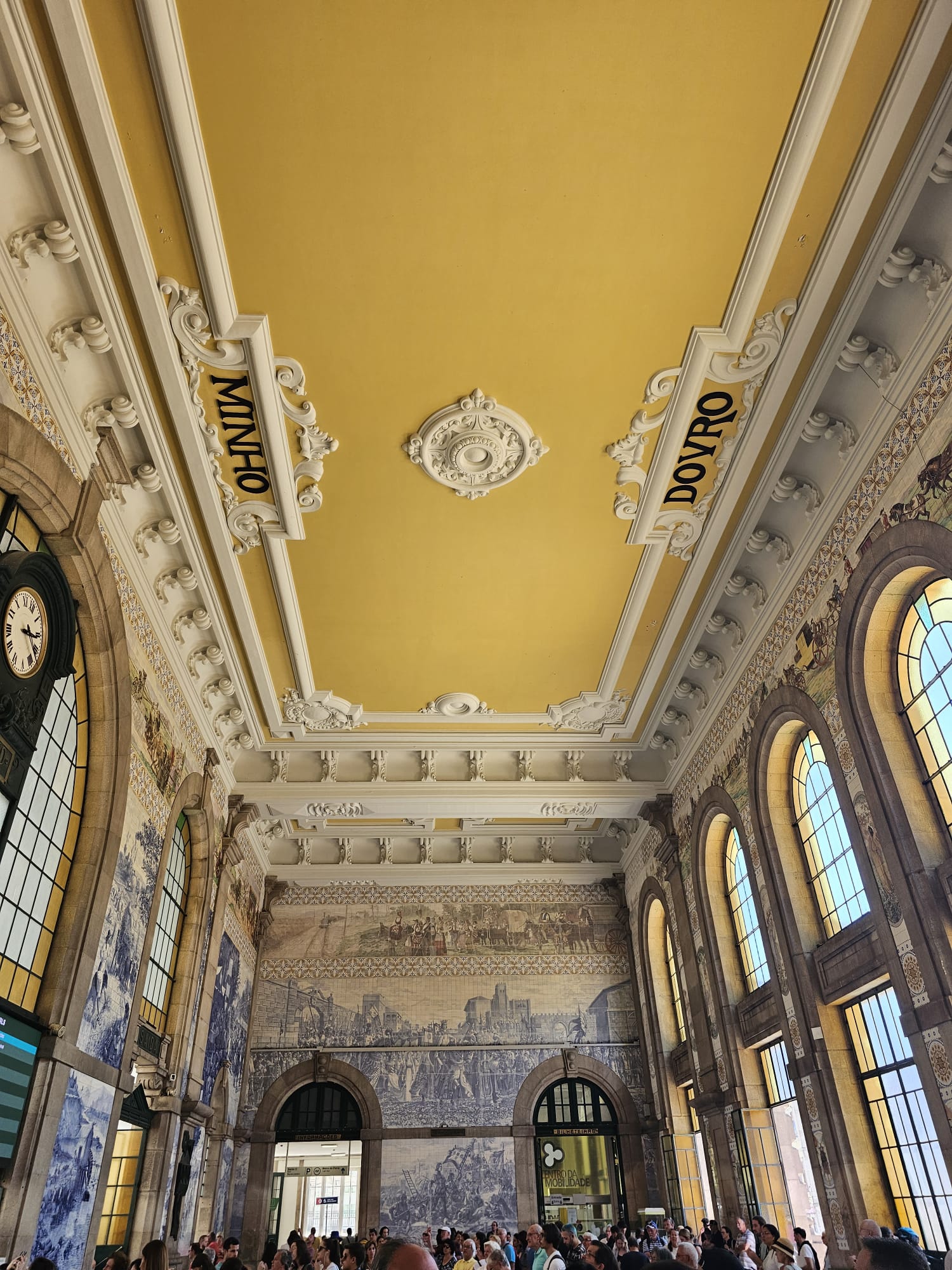 A grand interior of a train station features a high yellow ceiling with ornate white moldings and the words "Minho" and "Douro." The walls are adorned with intricate blue and white tile murals depicting historical scenes. Large arched windows allow natural light to flood the bustling space filled with people.