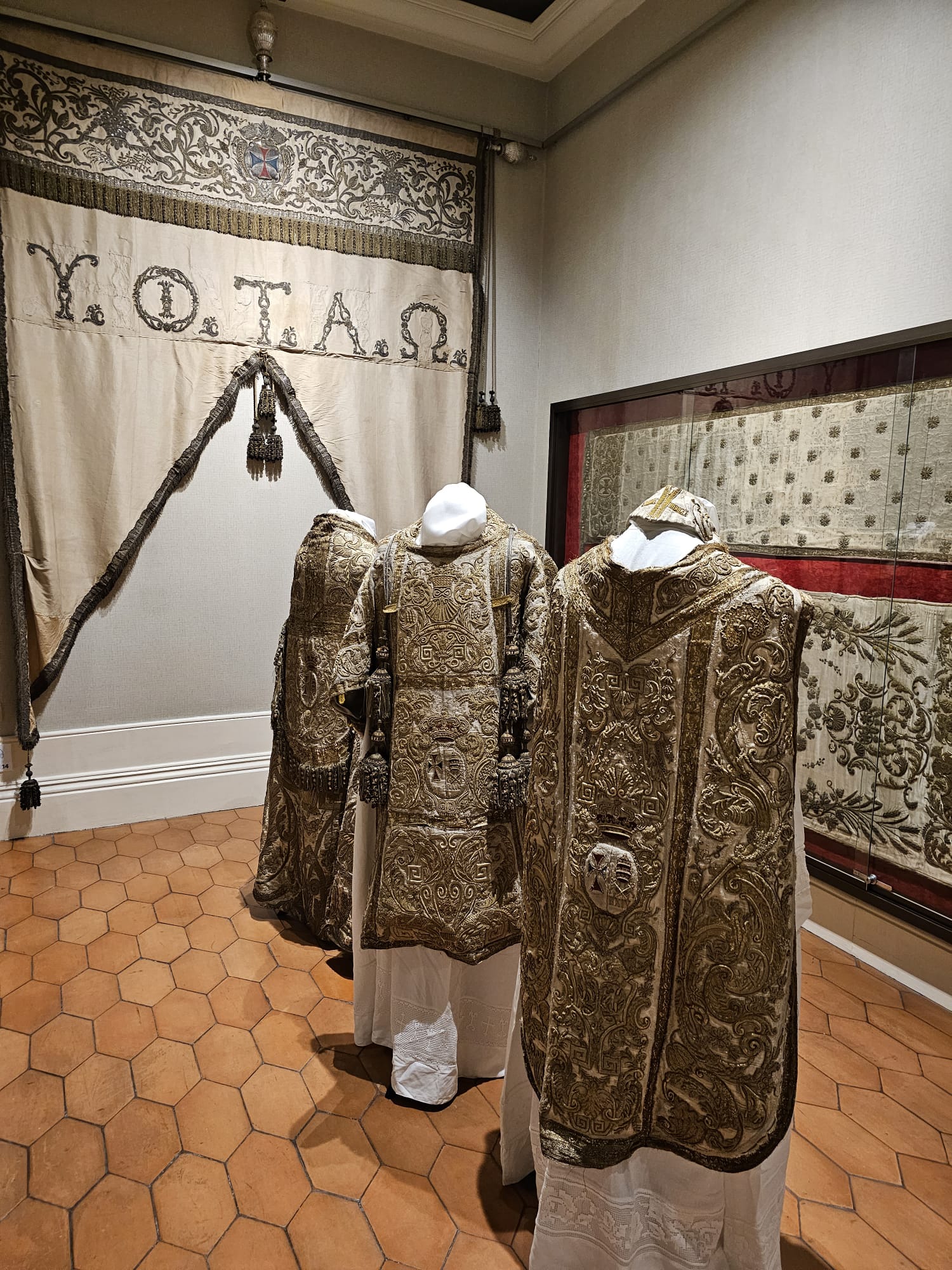 Three ornate, gold-embroidered vestments are displayed on mannequins in a museum setting, surrounded by intricate textiles and a decorative banner with Greek letters.