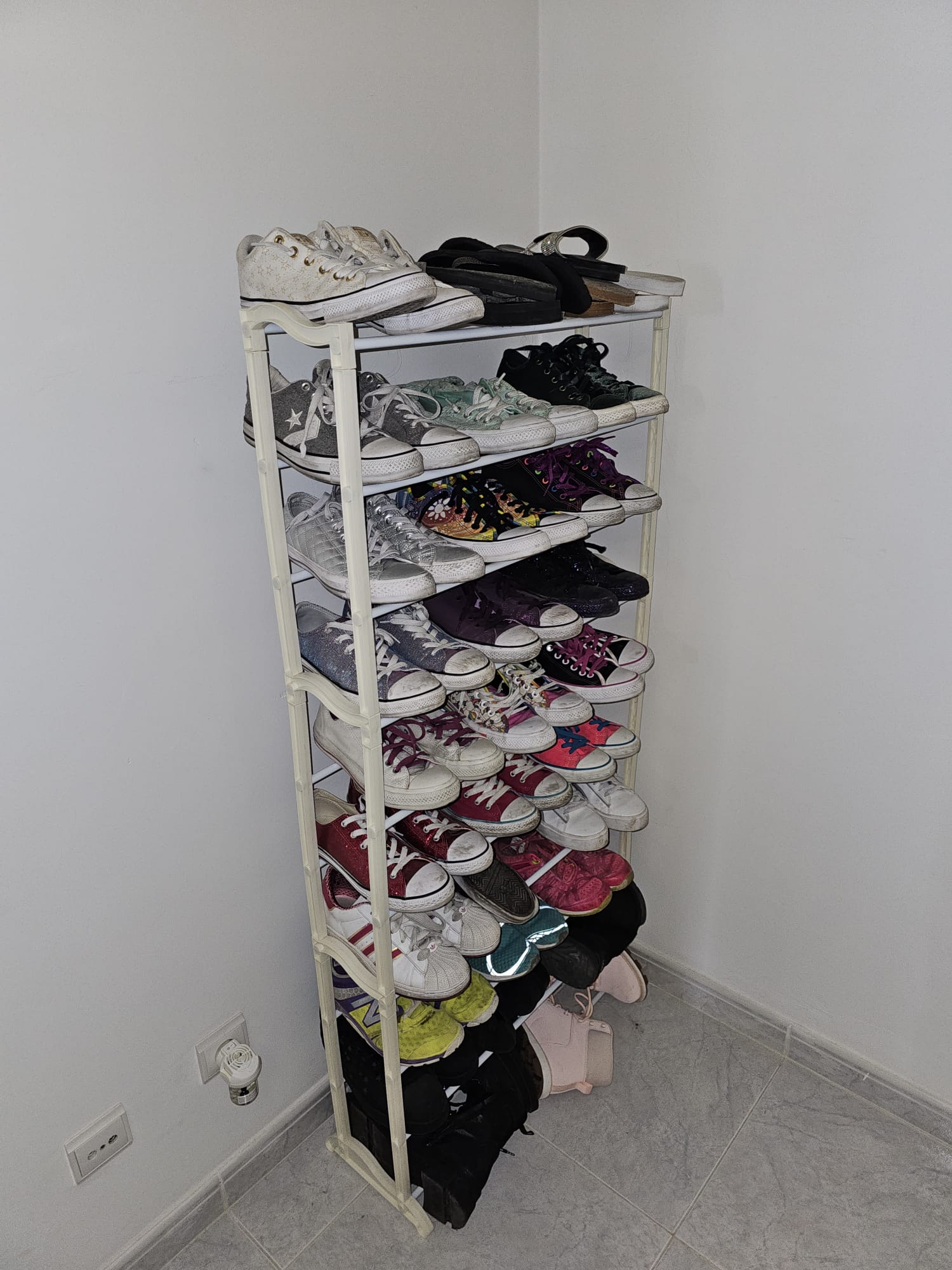 A tall shoe rack filled with various sneakers and shoes stands in the corner of a room. The shelves hold a mix of colorful and neutral-toned footwear, including Converse sneakers and flip-flops. The white walls and tiled floor create a clean, minimalist setting.