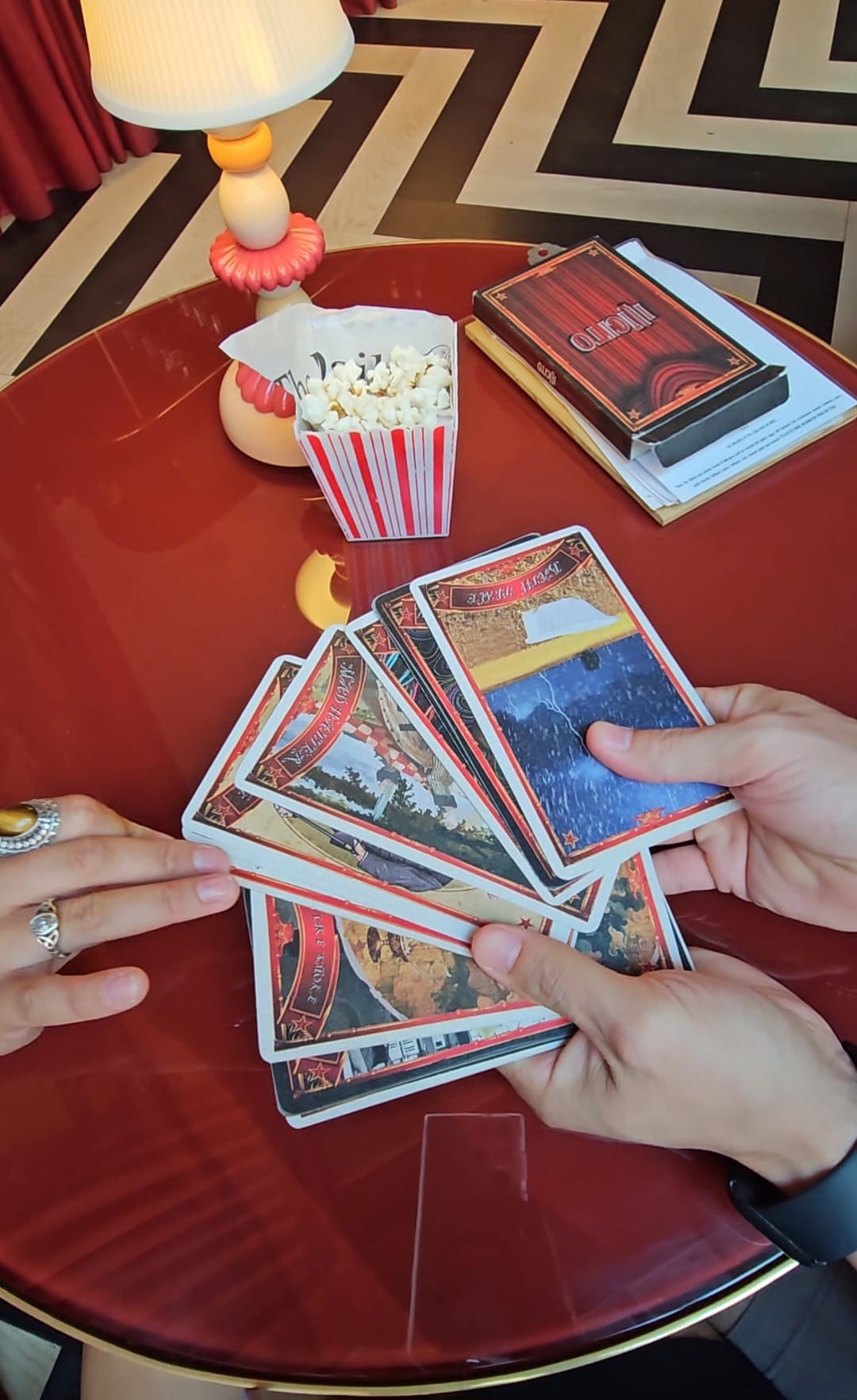 Two hands hold a spread of vintage-style tarot cards over a red table, with a small striped popcorn box and a decorative lamp nearby.