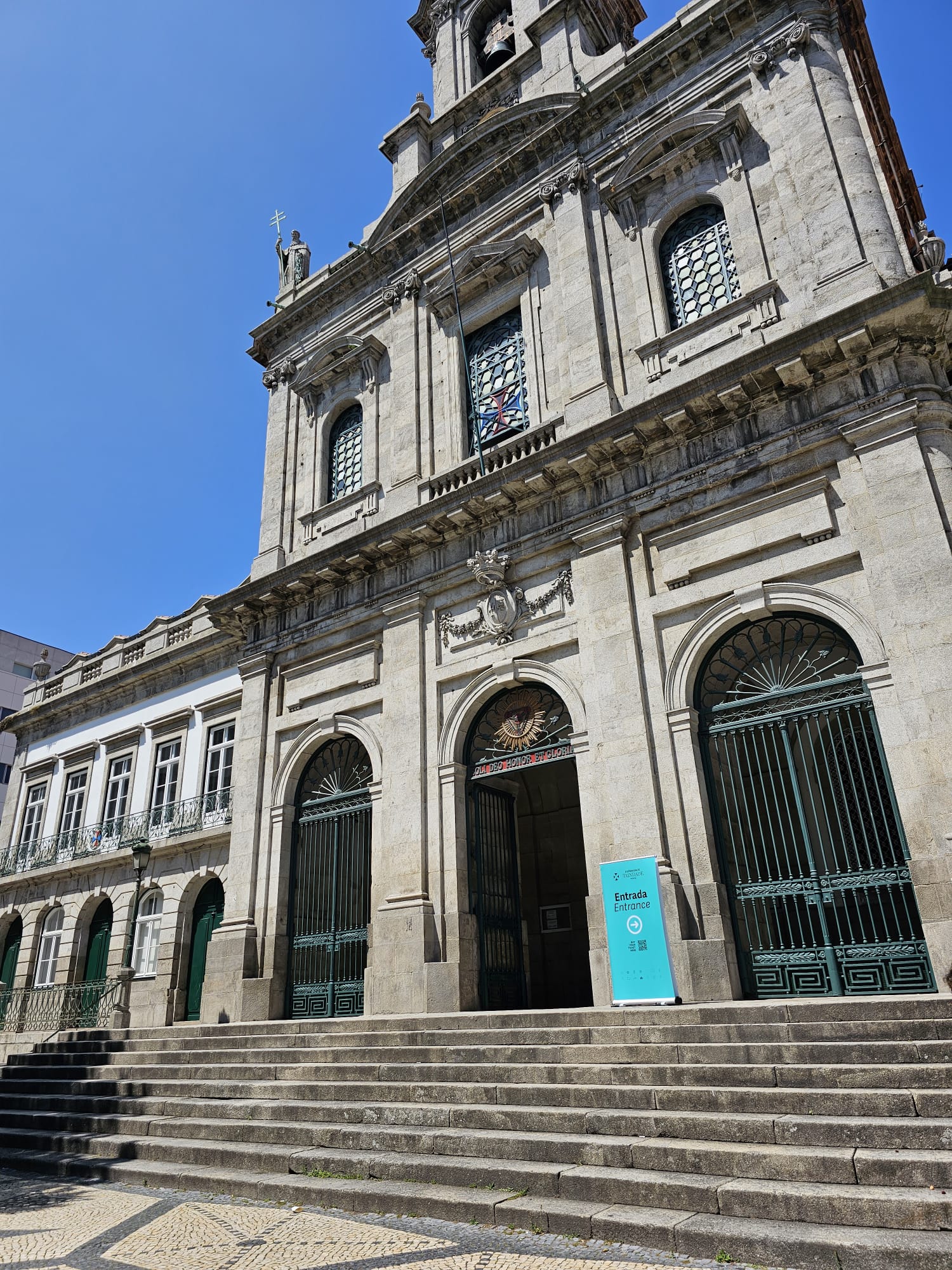 A grand stone building with ornate architectural details stands under a clear blue sky. The facade features arched windows with decorative grilles and a central entrance with large metal doors. A statue and a bell tower adorn the top. Stone steps lead up to the entrance, where a turquoise sign indicates "Entrance.".