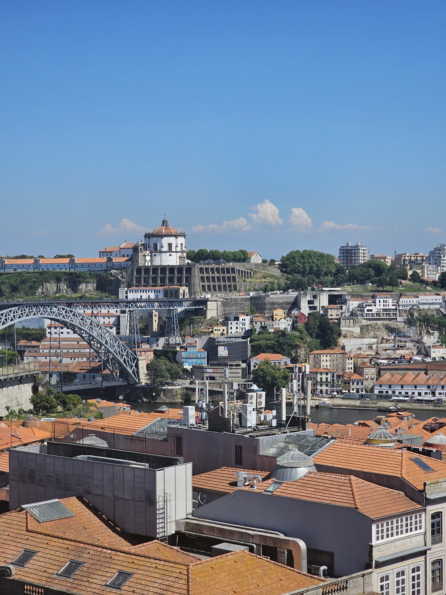 A panoramic view of a cityscape featuring a prominent arched bridge crossing a river, with a historic white building with a dome on a hill in the background. The foreground shows a cluster of buildings with red-tiled roofs under a clear blue sky.