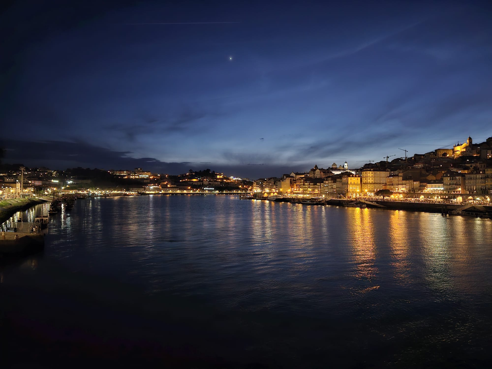 A picturesque evening view of a riverside cityscape, with buildings warmly illuminated along the water's edge. The sky is a deep blue, with a faint star visible, and the reflections of the lights shimmer on the calm river.