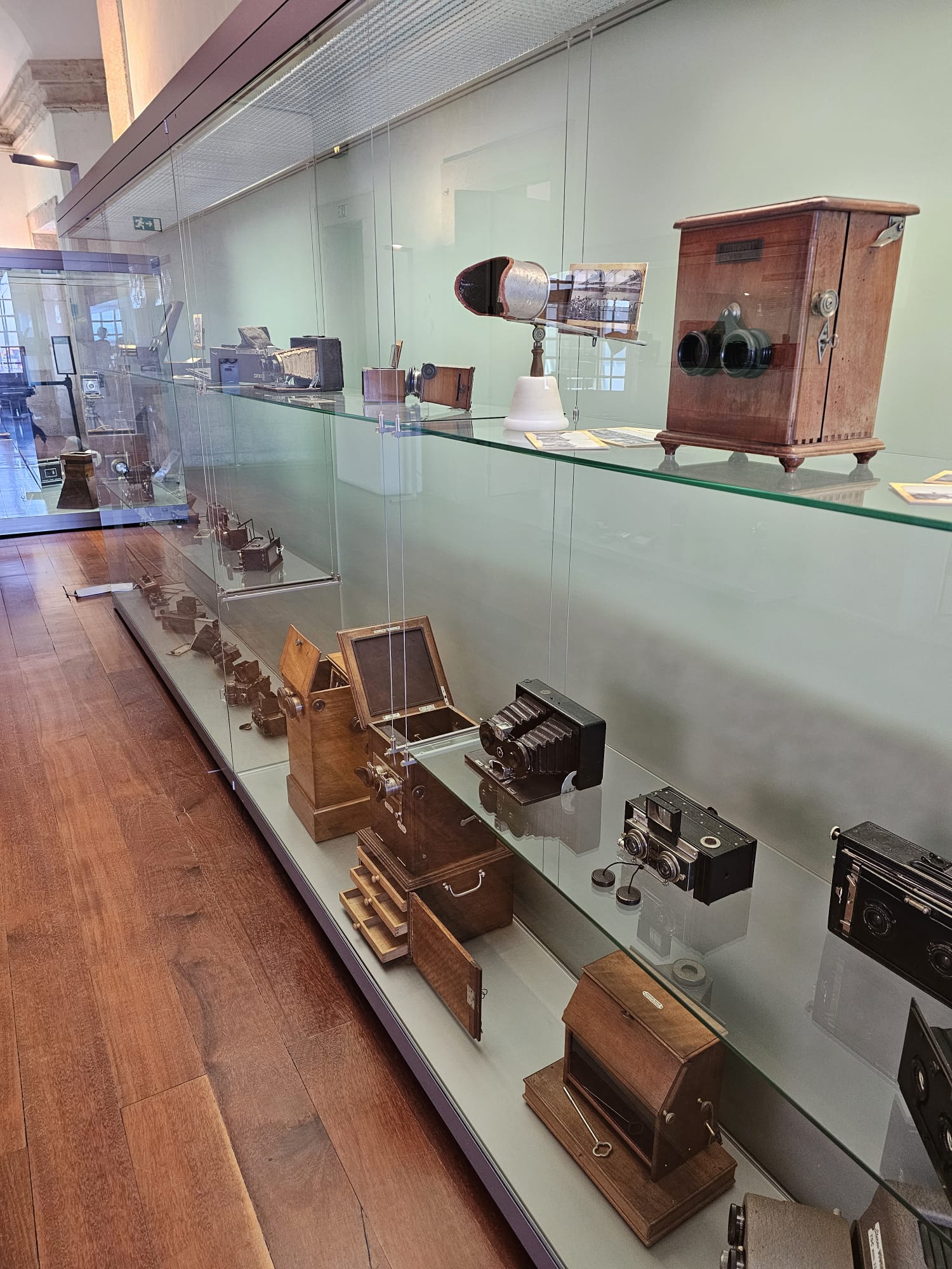 A glass display case in a museum showcases a collection of vintage cameras and optical devices. The items, primarily made of wood and metal, vary in size and design, lined up on two shelves. The floor is polished wood, and the setting is bright and spacious.