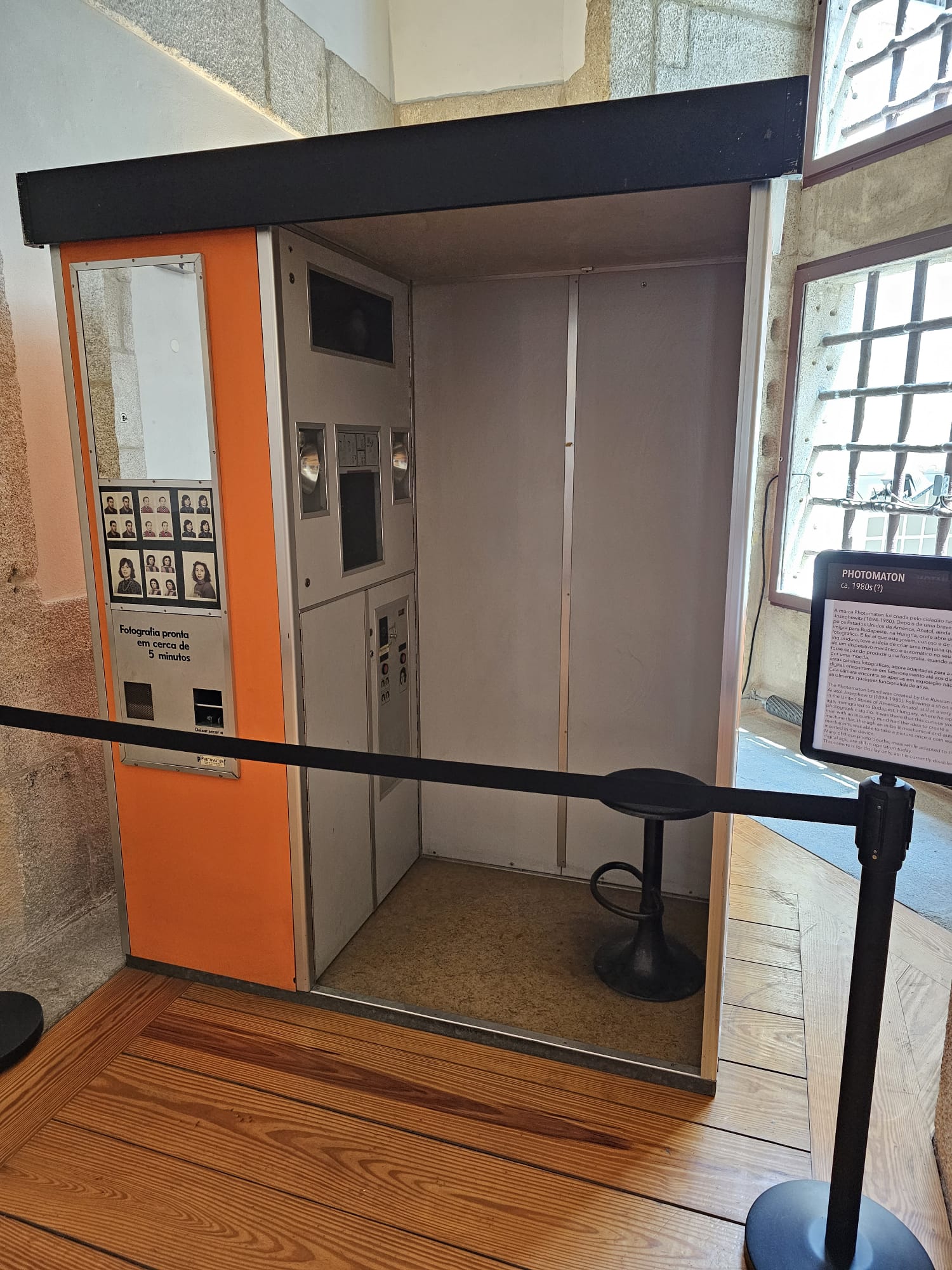 A vintage photo booth with an orange and gray exterior, featuring a small stool inside, is displayed in a museum setting. A black barrier cordon surrounds it, and a sign with information is positioned nearby.