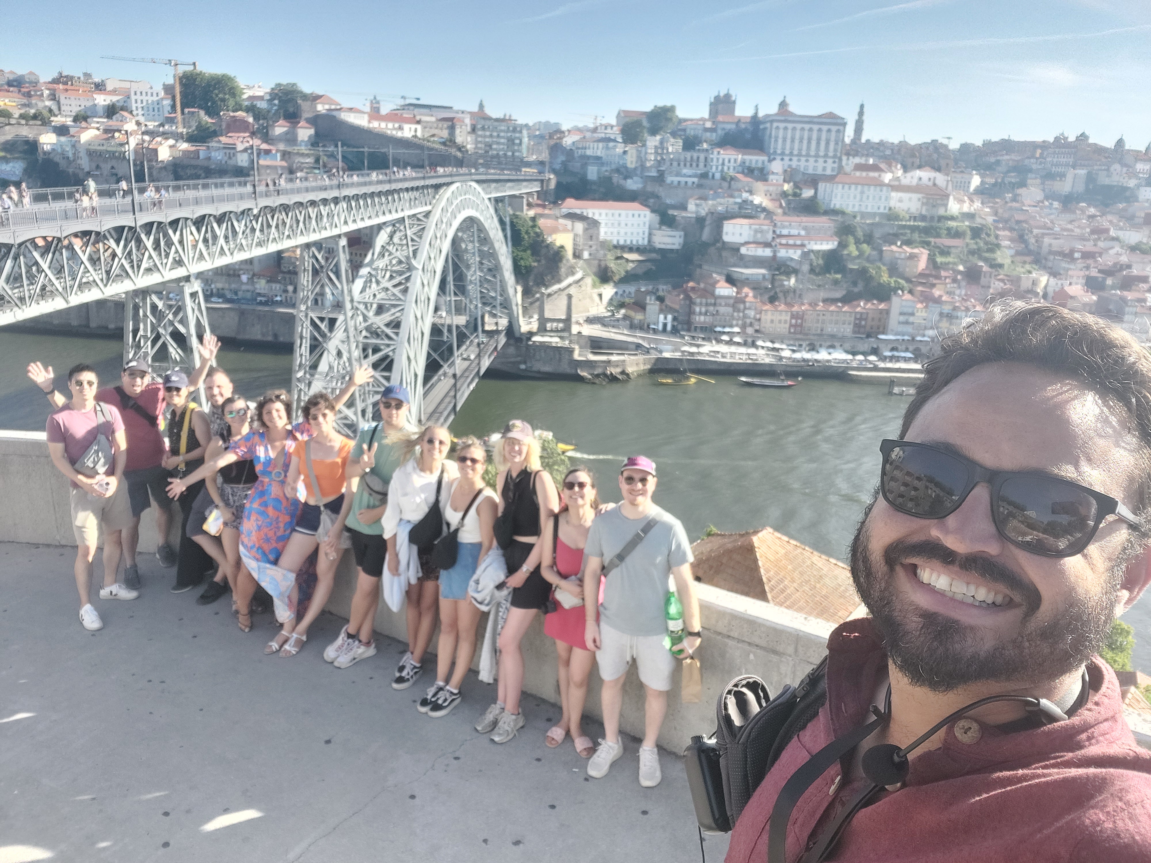 A group of people poses for a cheerful selfie on a sunny day, with a large, arched metal bridge and a scenic cityscape in the background. The river below reflects the clear blue sky, and the city buildings rise on a hillside, creating a picturesque view.