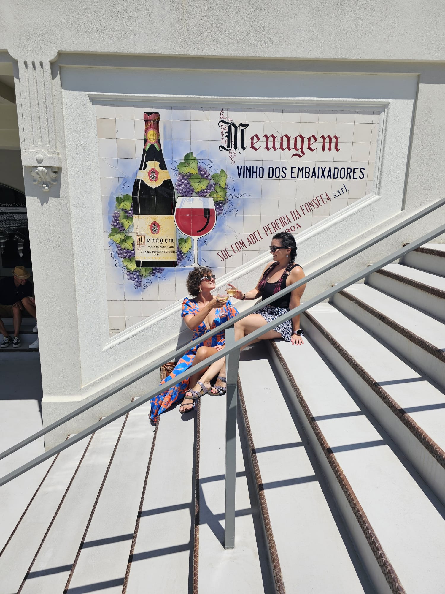 Two women sit on outdoor steps, sharing a drink and smiling. Behind them is a colorful mural featuring a wine bottle and glass, with the words "Menagem Vinho dos Embaixadores." The scene is sunny and relaxed.