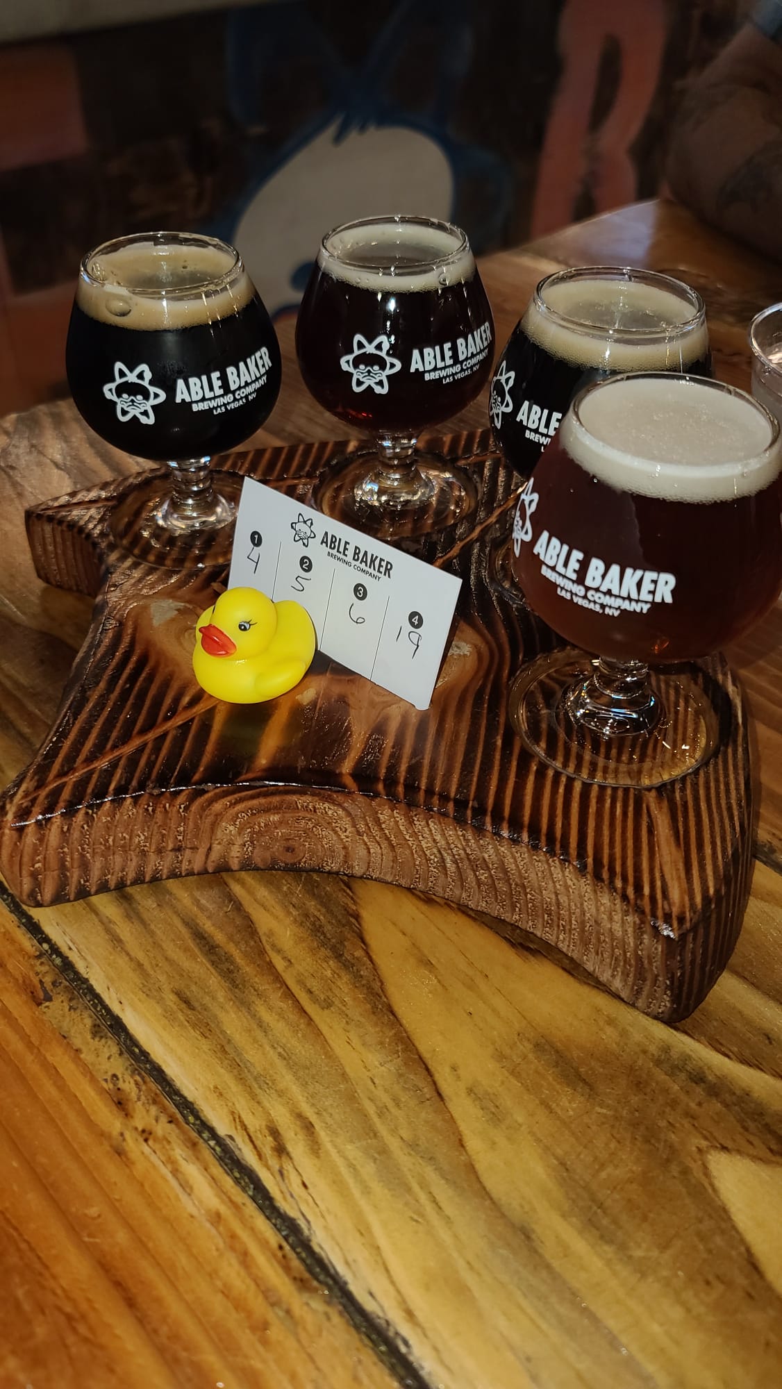 A wooden tray holds four small glasses of dark beer, each labeled with "Able Baker Brewing Company." A yellow rubber duck and a tasting card are placed on the tray, set on a rustic wooden table.