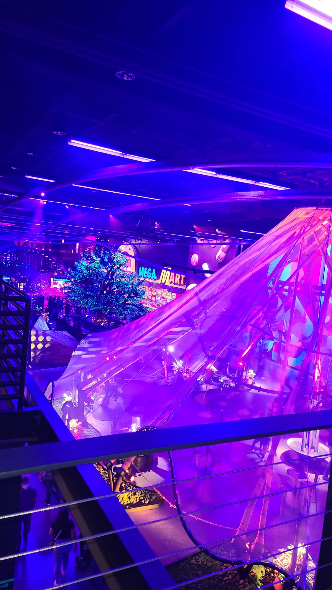 A vibrant indoor scene bathed in purple and blue neon lights, featuring a large, illuminated artificial tree and a sign reading "MEGA MART." The space is filled with colorful displays and structures, creating a futuristic, immersive atmosphere.