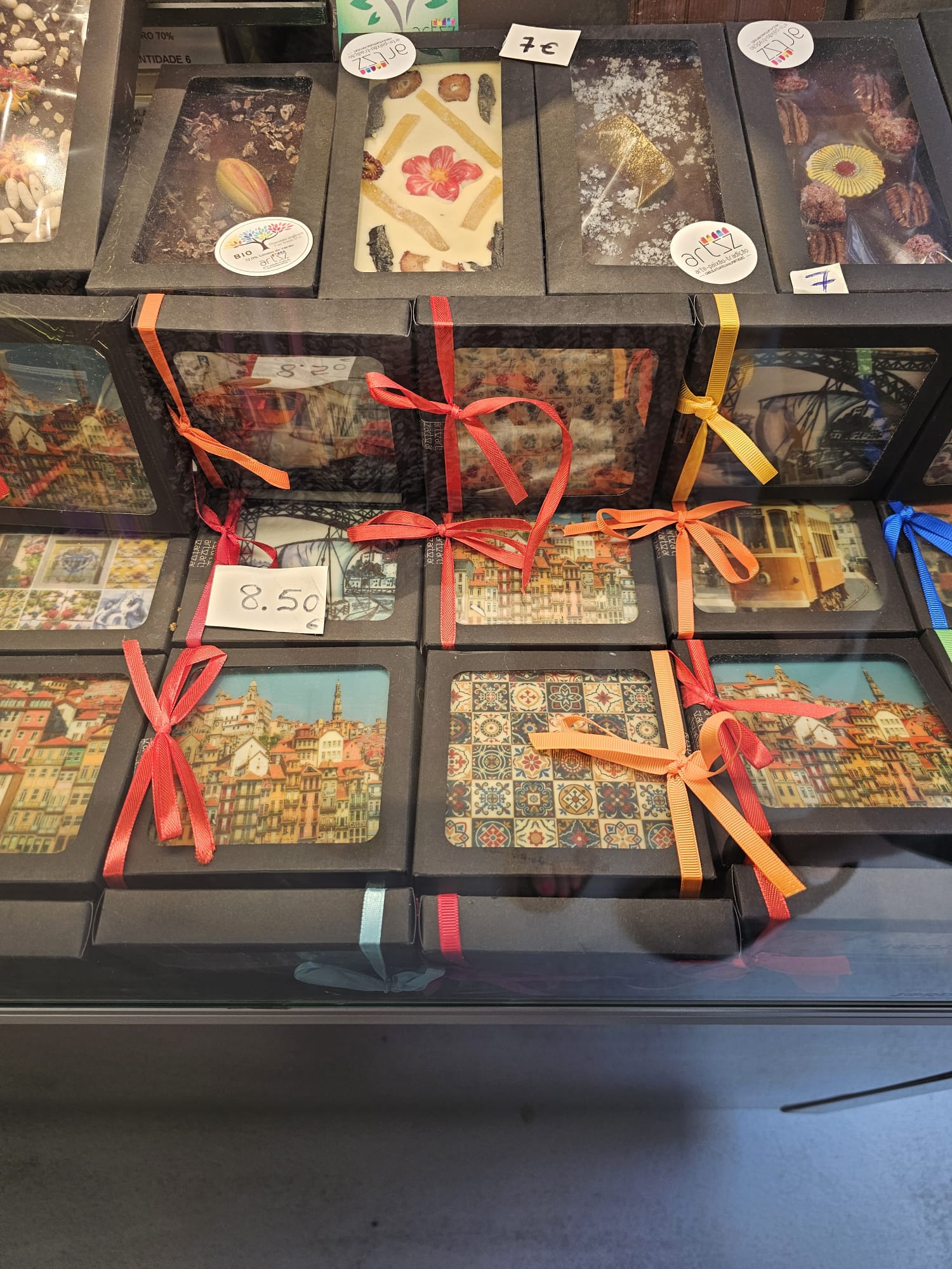 A display of chocolate bars in black boxes with transparent windows, showcasing various toppings like dried flowers and nuts. Each box is adorned with colorful ribbons, including red, orange, and yellow, and some have price tags like €7 and €8.50. The packaging features vibrant images of cityscapes and intricate tile patterns.