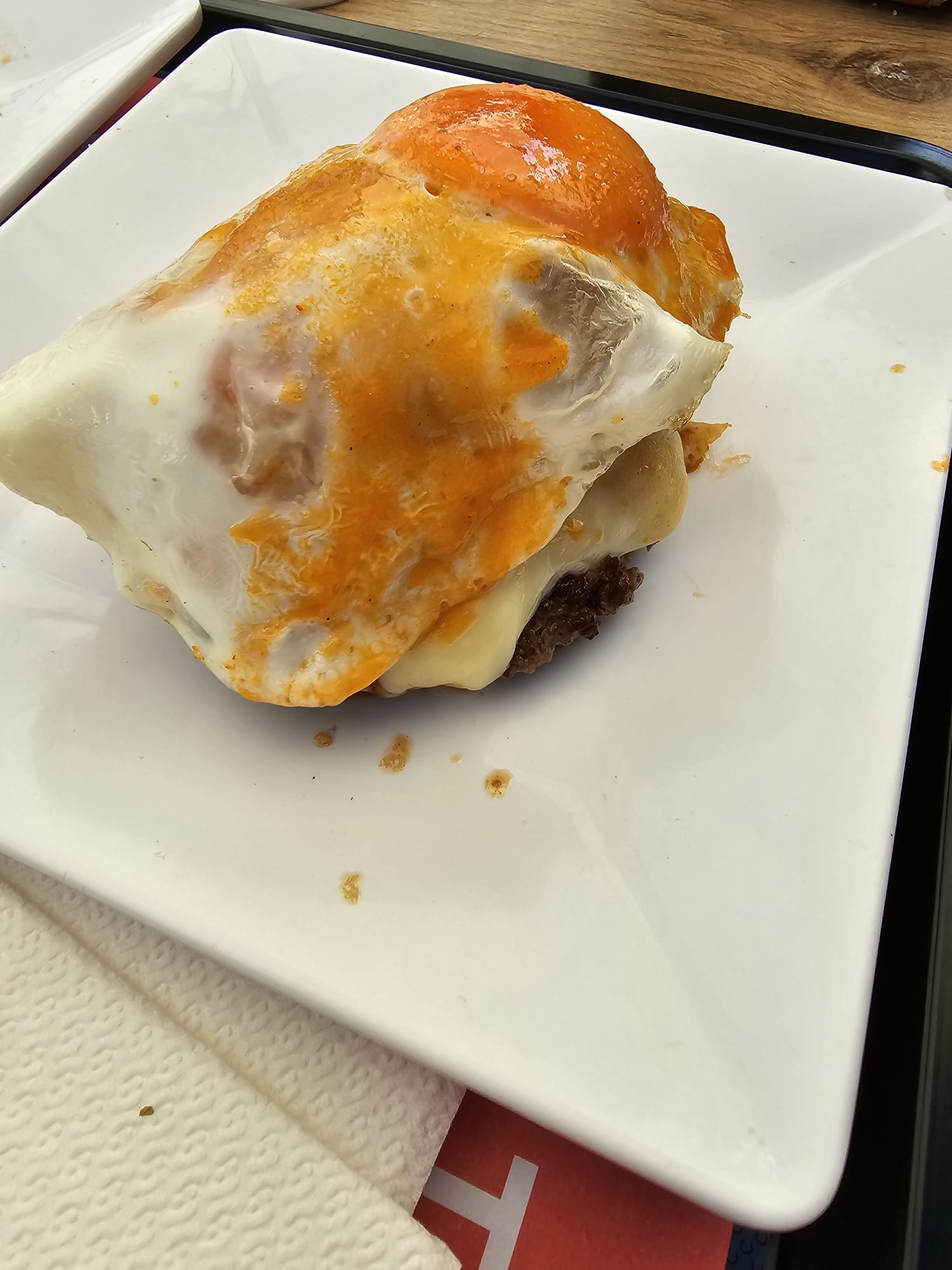 A hamburger topped with a fried egg and melted cheese sits on a white square plate. The egg's yolk is bright orange and slightly runny, adding a rich texture to the dish.