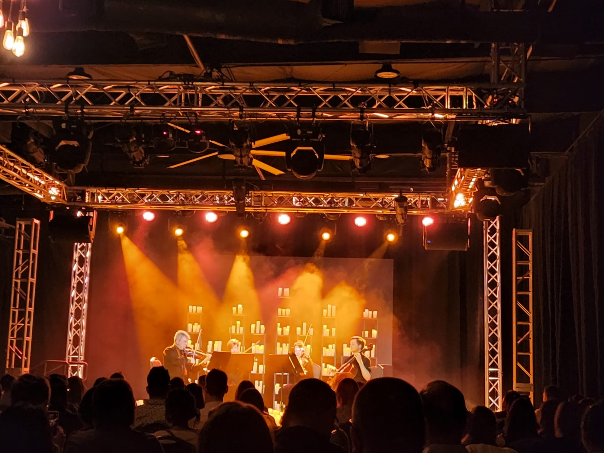 A quartet of musicians performs on a dimly lit stage, surrounded by a warm glow from numerous candles arranged on shelves in the background. The audience is seated in the foreground, attentively watching the performance. The stage is framed by metal trusses and illuminated by orange and red spotlights.
