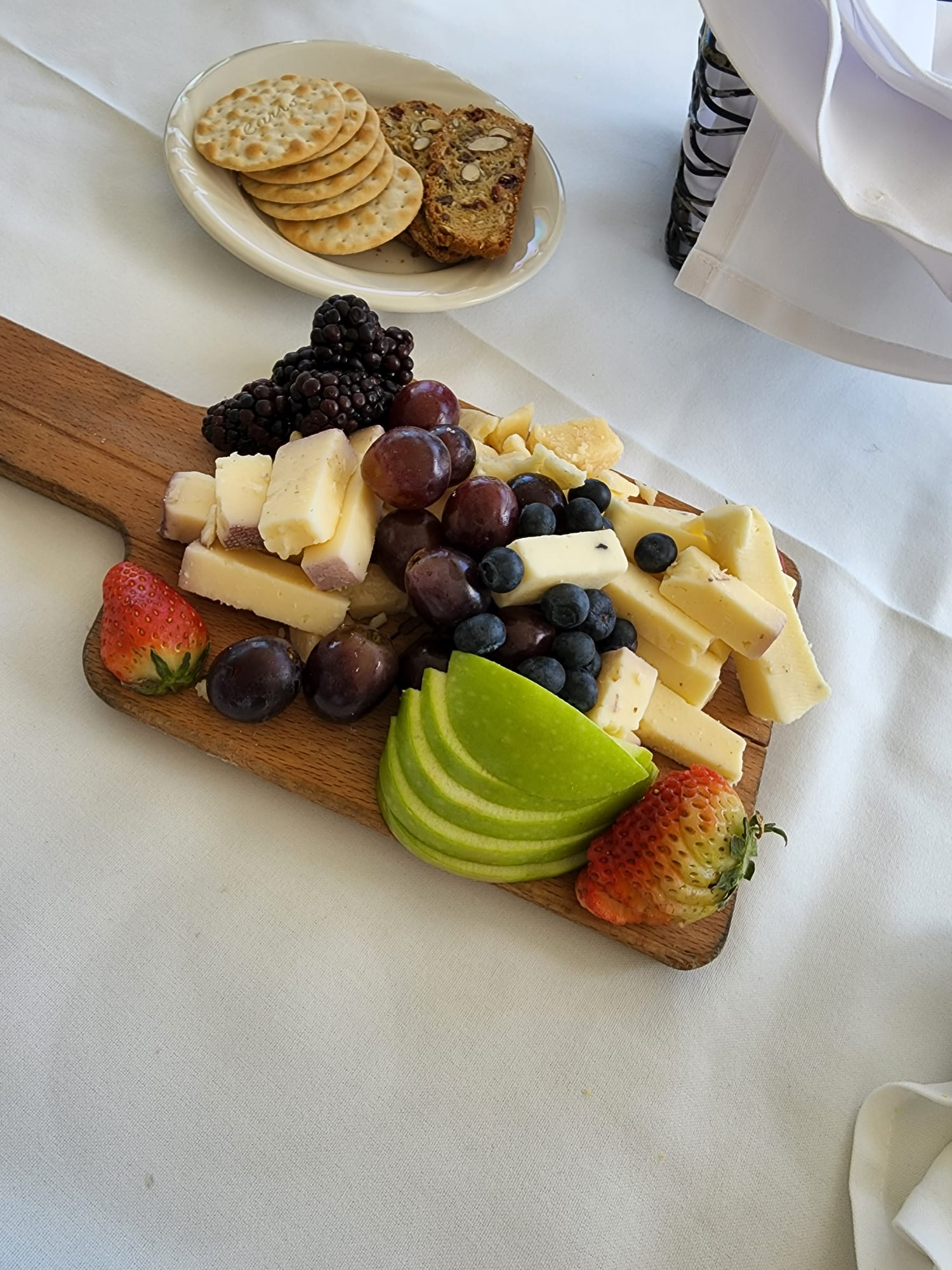A wooden board displays a selection of cheeses, grapes, blackberries, blueberries, and sliced green apples, with strawberries at the edges. Nearby, a plate holds assorted crackers and nut-studded bread, all set on a white tablecloth.
