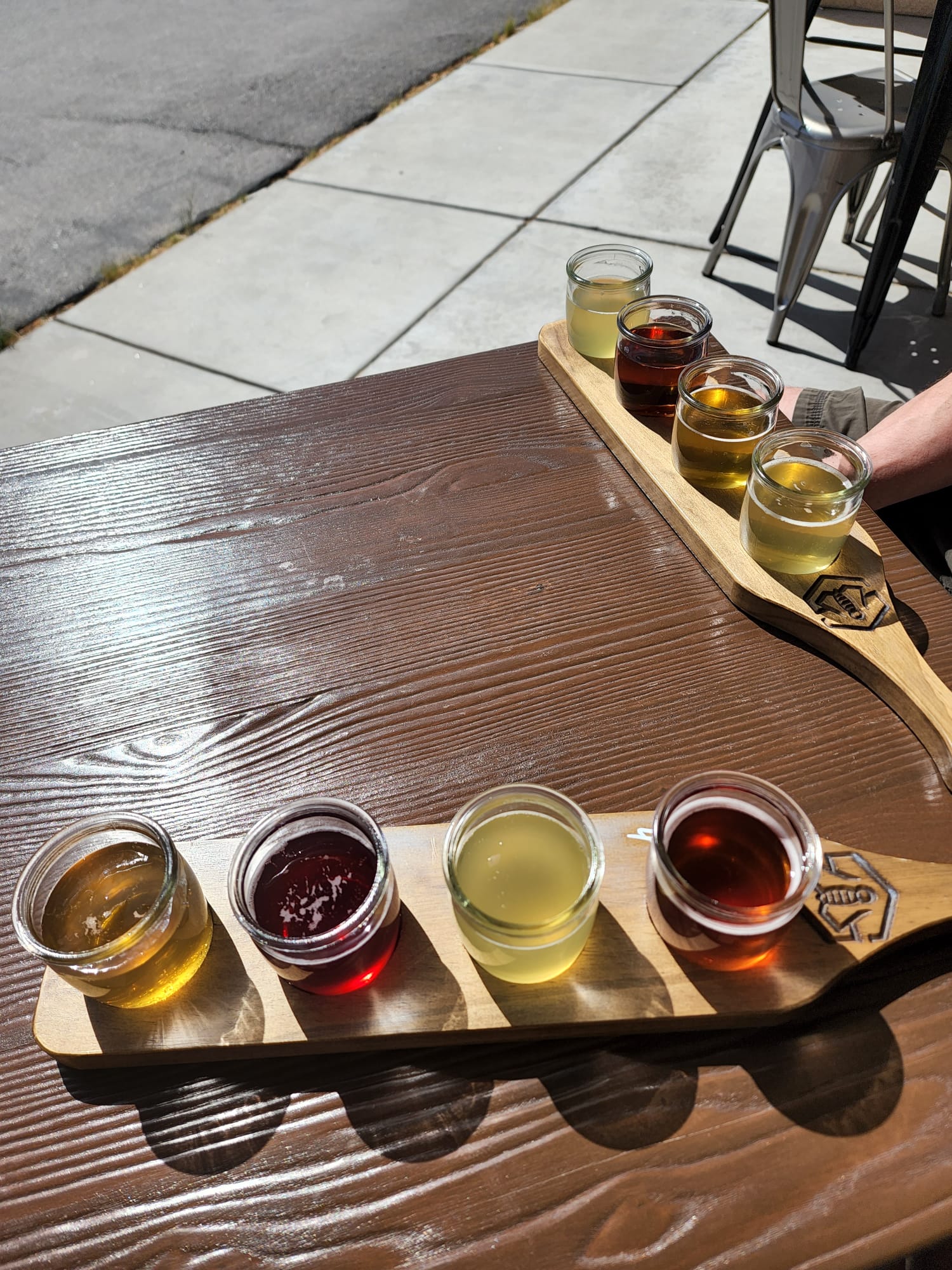 A wooden table holds two flight boards, each with four small glass jars filled with different colored beverages, ranging from amber to deep red. The scene is set outdoors, with a sidewalk visible in the background. Sunlight casts shadows, highlighting the vibrant colors of the drinks.