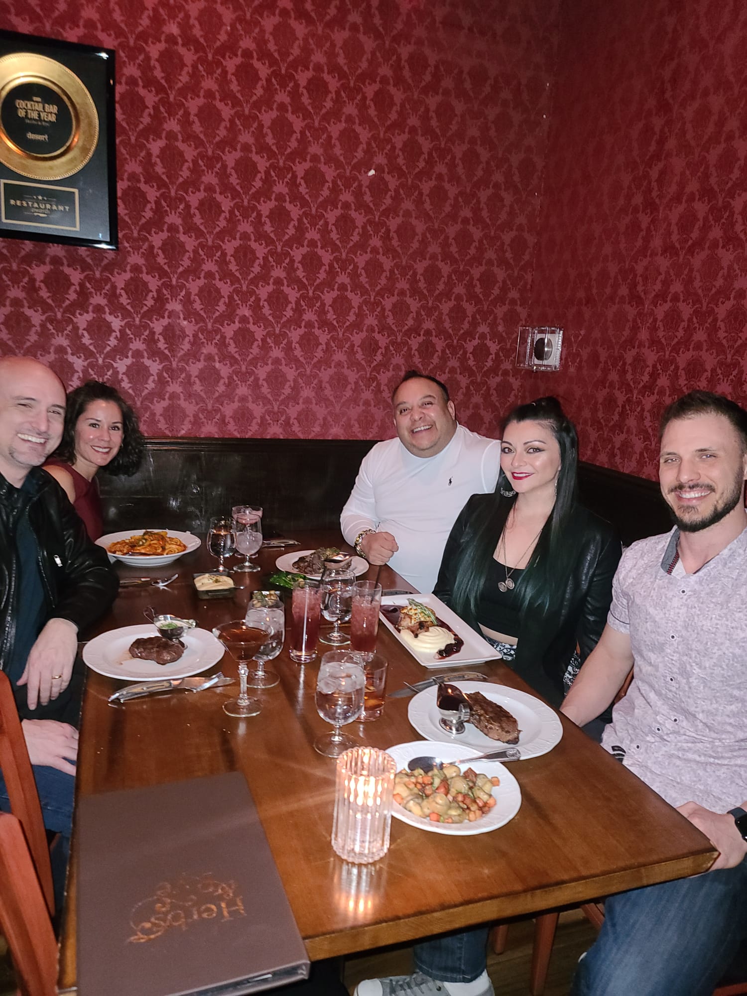 A group of five people is seated around a wooden table in a restaurant with red patterned walls. They are enjoying a meal with plates of steak, pasta, and assorted dishes in front of them. A candle and drinks are also on the table, and a plaque on the wall reads "Cocktail Bar of the Year.".