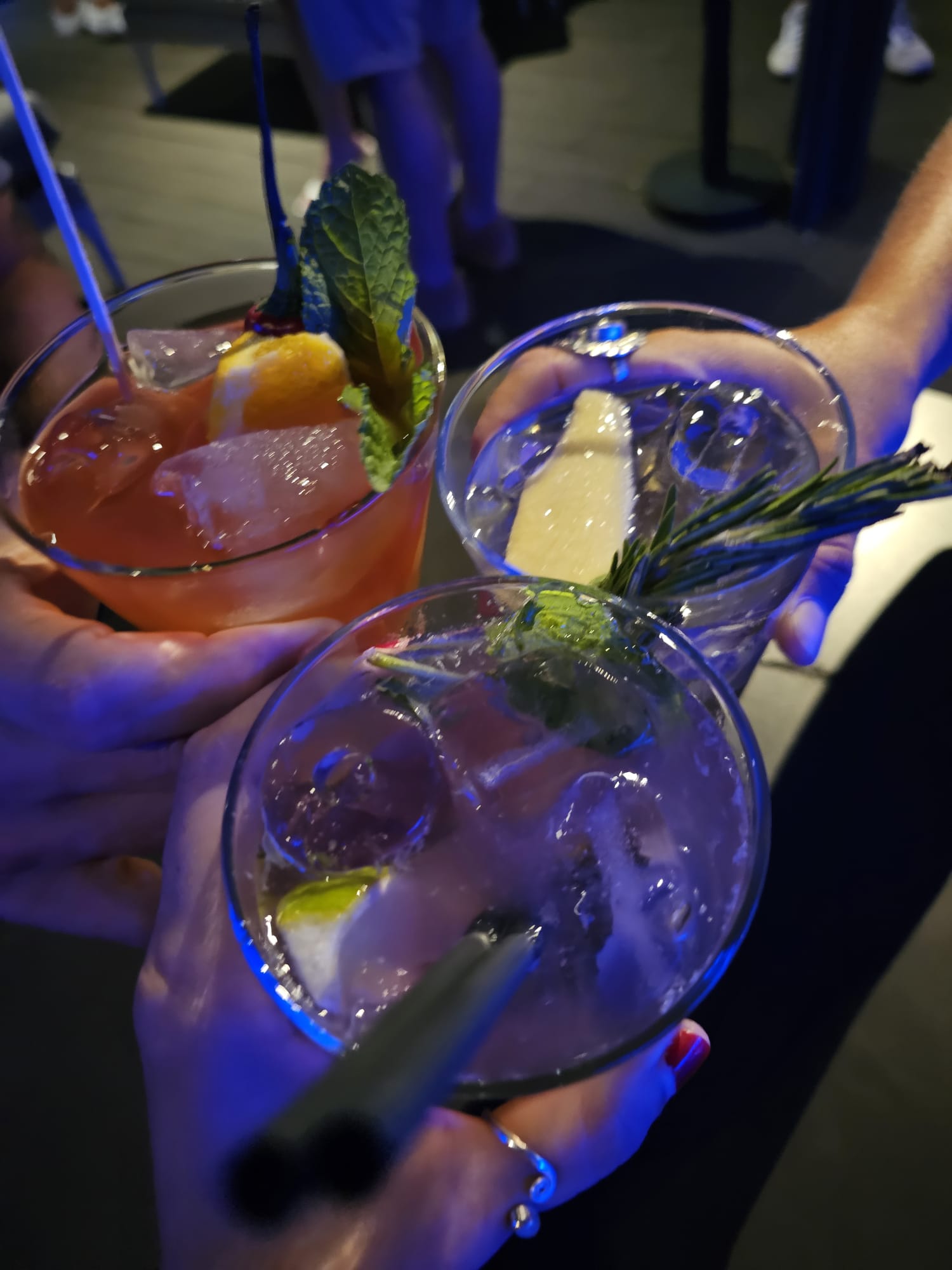 Three hands hold glasses of colorful cocktails with ice, garnished with mint leaves and citrus slices, under ambient blue lighting in a social setting.