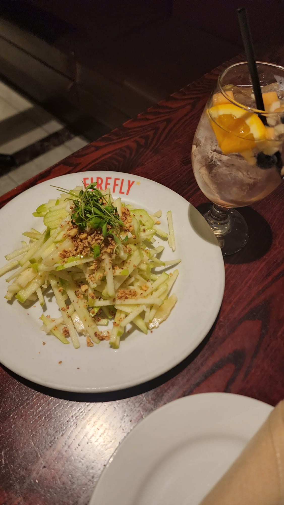 A fresh green apple salad topped with chopped nuts and greens is served on a white plate with "FIREFLY" written on it, accompanied by a refreshing drink garnished with orange slices and berries in a glass with a black straw, all set on a dark wooden table.