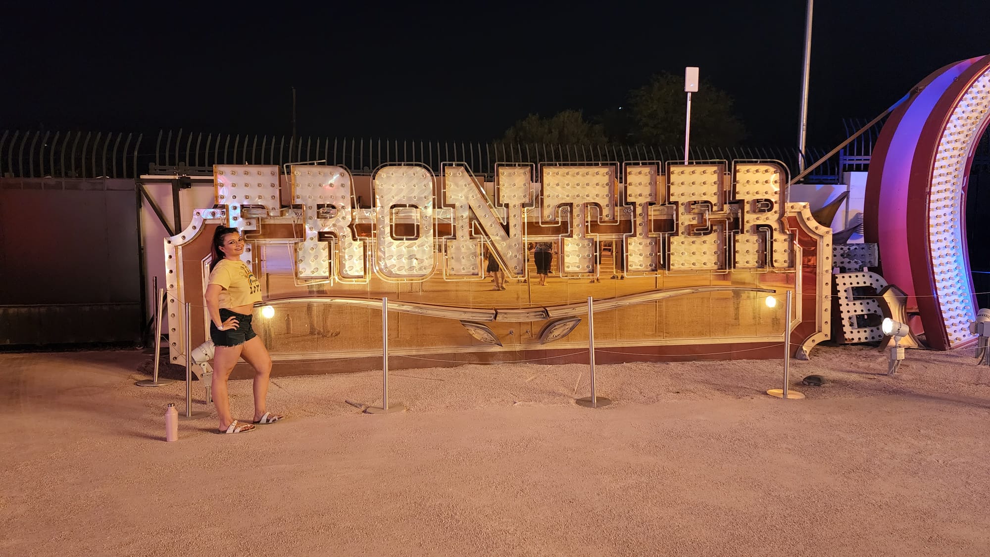 A person in a yellow shirt and shorts poses confidently next to a large, illuminated "FRONTIER" sign at night. The sign has a vintage style with numerous light bulbs, and the scene is set outdoors on a gravel surface.