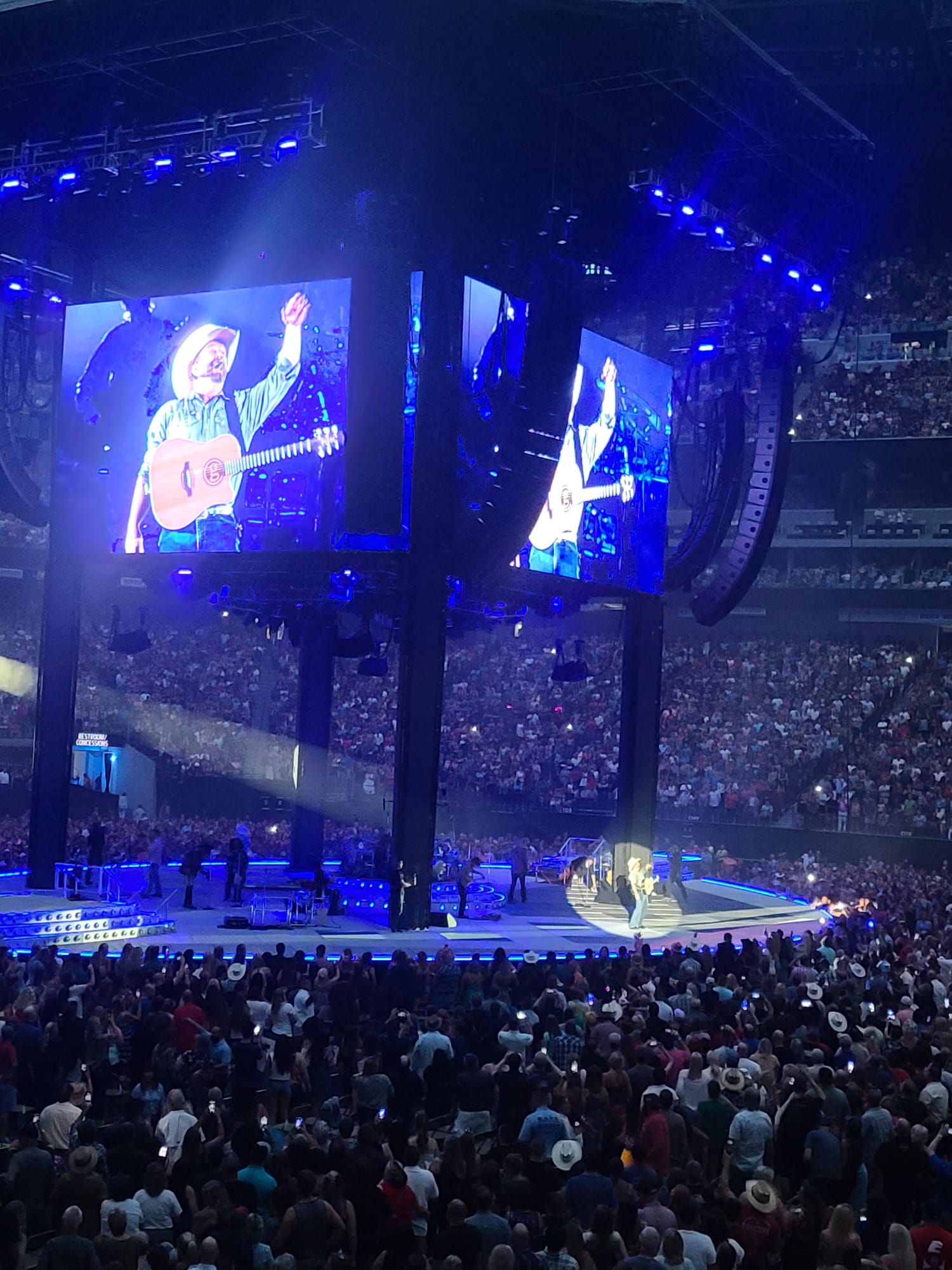 A lively concert scene with a performer in a cowboy hat and guitar on a brightly lit stage, surrounded by a large, enthusiastic crowd. The performer is shown on a big screen above the stage, which is illuminated with blue and purple lights, enhancing the vibrant atmosphere.