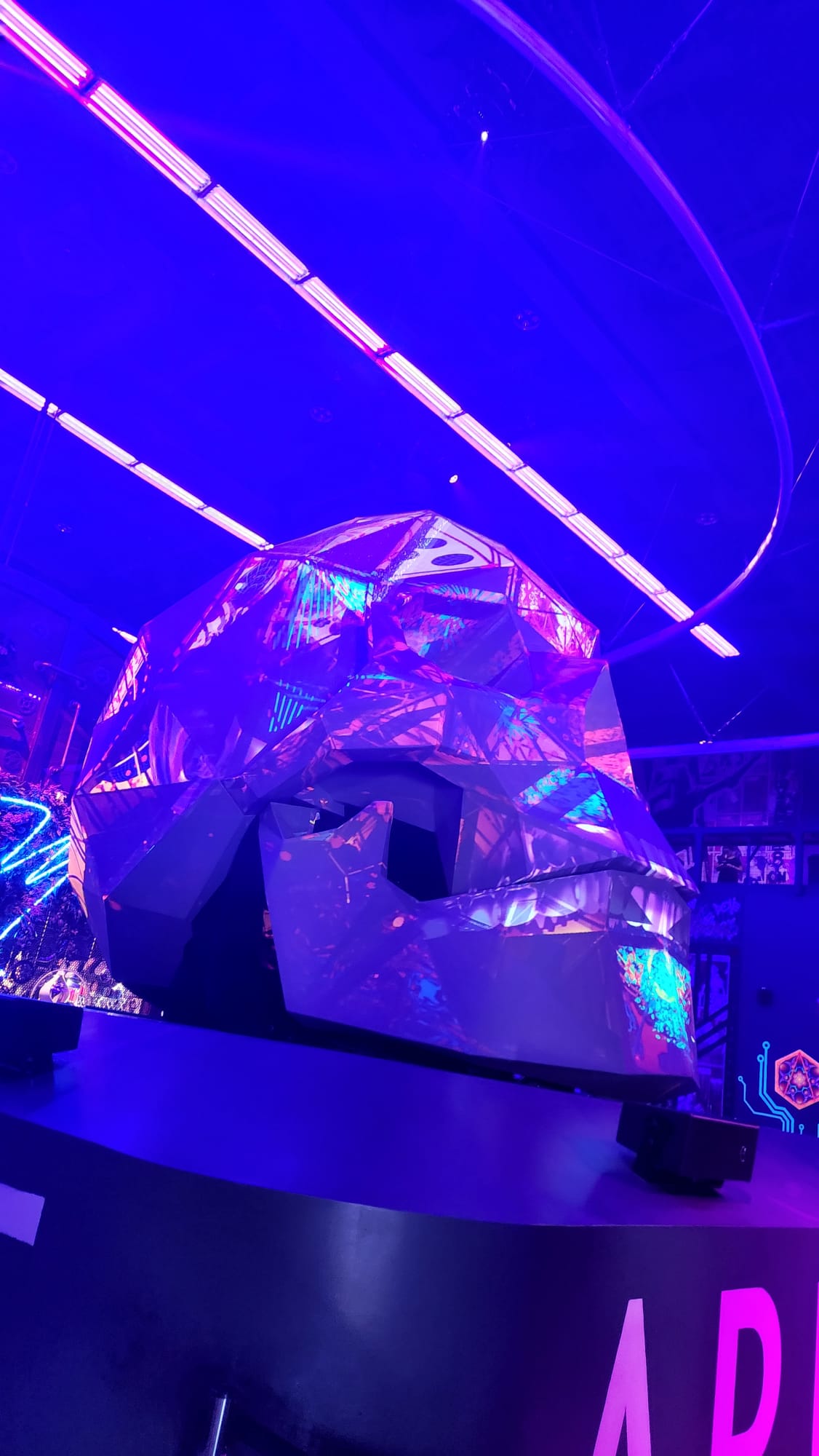 A futuristic, faceted sculpture illuminated in vibrant purple and blue lights sits on display in a dimly lit space, surrounded by neon accents and reflective surfaces.