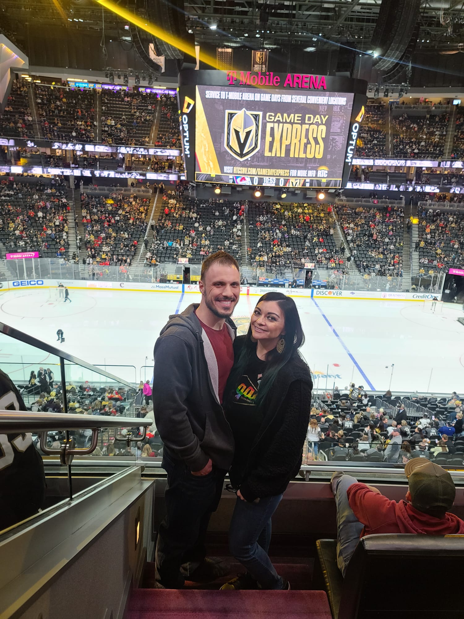 A couple stands smiling in a hockey arena, with the ice rink and a large scoreboard in the background. The arena is filled with spectators, and the scoreboard displays "Game Day Express." They are positioned near a railing, dressed casually in jackets and jeans.