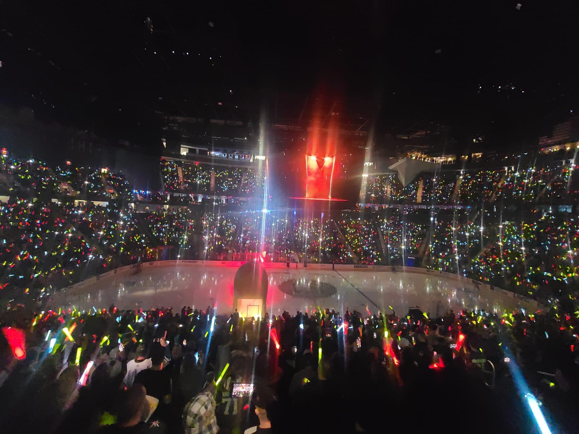 A vibrant hockey arena is filled with spectators waving colorful light sticks, creating a dazzling display around the ice rink. The arena is dimly lit, with bright lights and a large screen above the rink adding to the lively atmosphere.