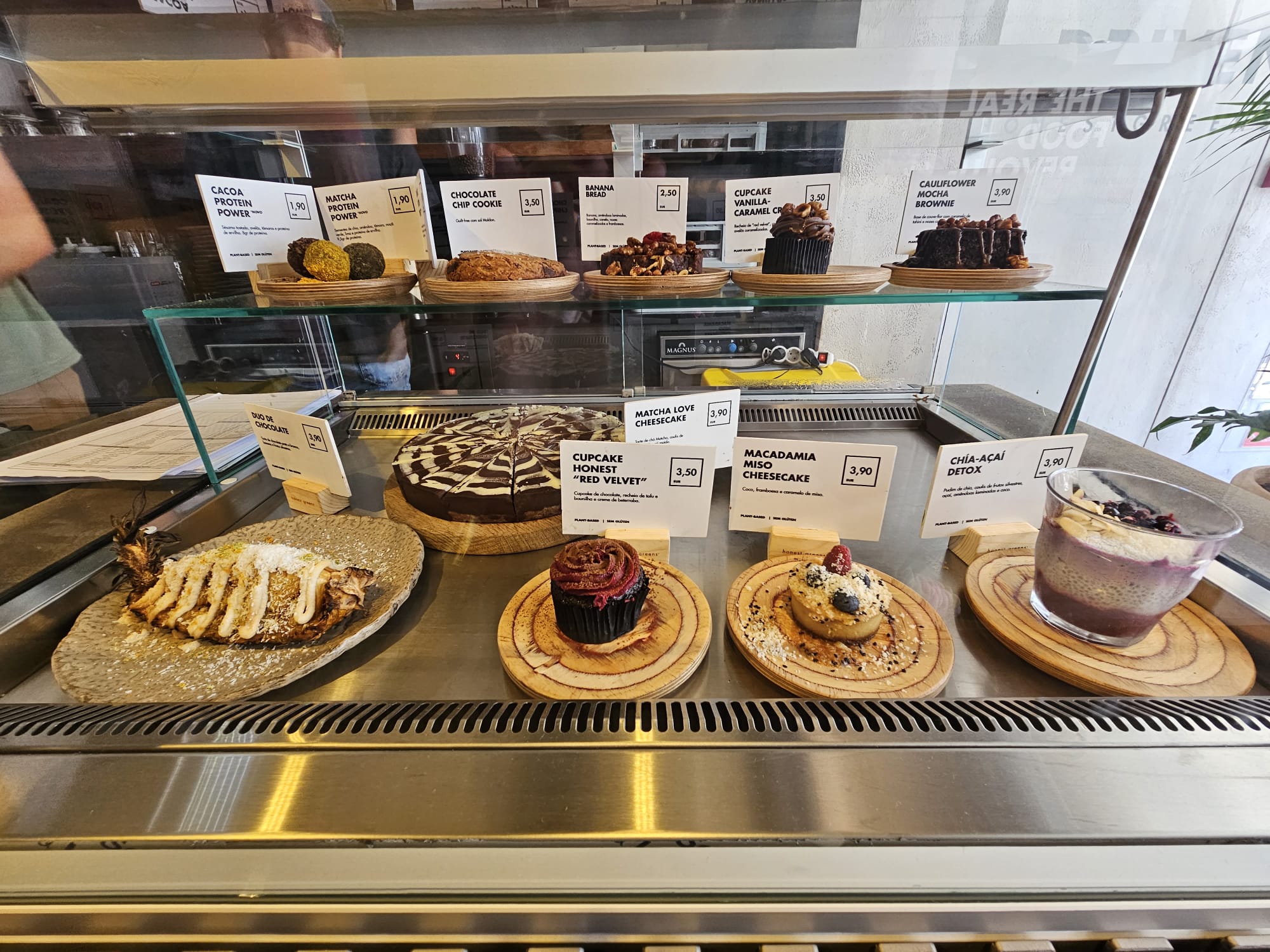 A display case filled with a variety of desserts, each labeled with a name and price. On the top shelf are items like a chocolate chip cookie and a cauliflower mocha brownie. The bottom shelf features a decorated cake, a red velvet cupcake, a macadamia miso cheesecake, and a chia-açaí detox bowl. The desserts are attractively arranged on wooden plates.