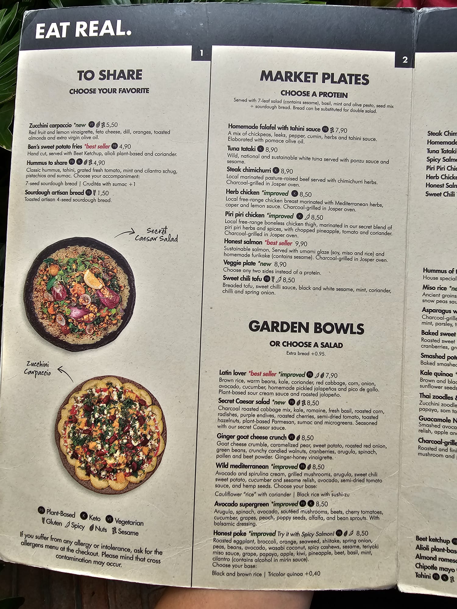 A menu titled "Eat Real" featuring sections for "To Share," "Market Plates," and "Garden Bowls." The "To Share" section includes options like Zucchini Carpaccio and Ben's Sweet Potato Fries. "Market Plates" offers proteins like Homemade Falafel and Tuna Tataki. "Garden Bowls" includes the Latin Lover and Secret Caesar Salad. Each item lists ingredients and prices, with symbols indicating dietary options such as plant-based.