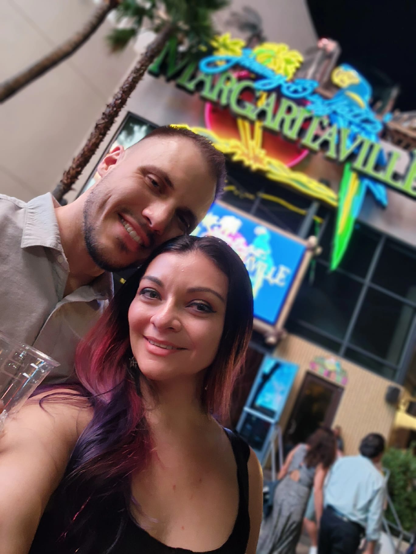 A smiling couple takes a selfie in front of a Margaritaville restaurant, with a colorful neon sign glowing above them. The man holds a clear plastic cup, and the woman's long hair has vibrant red and purple hues.