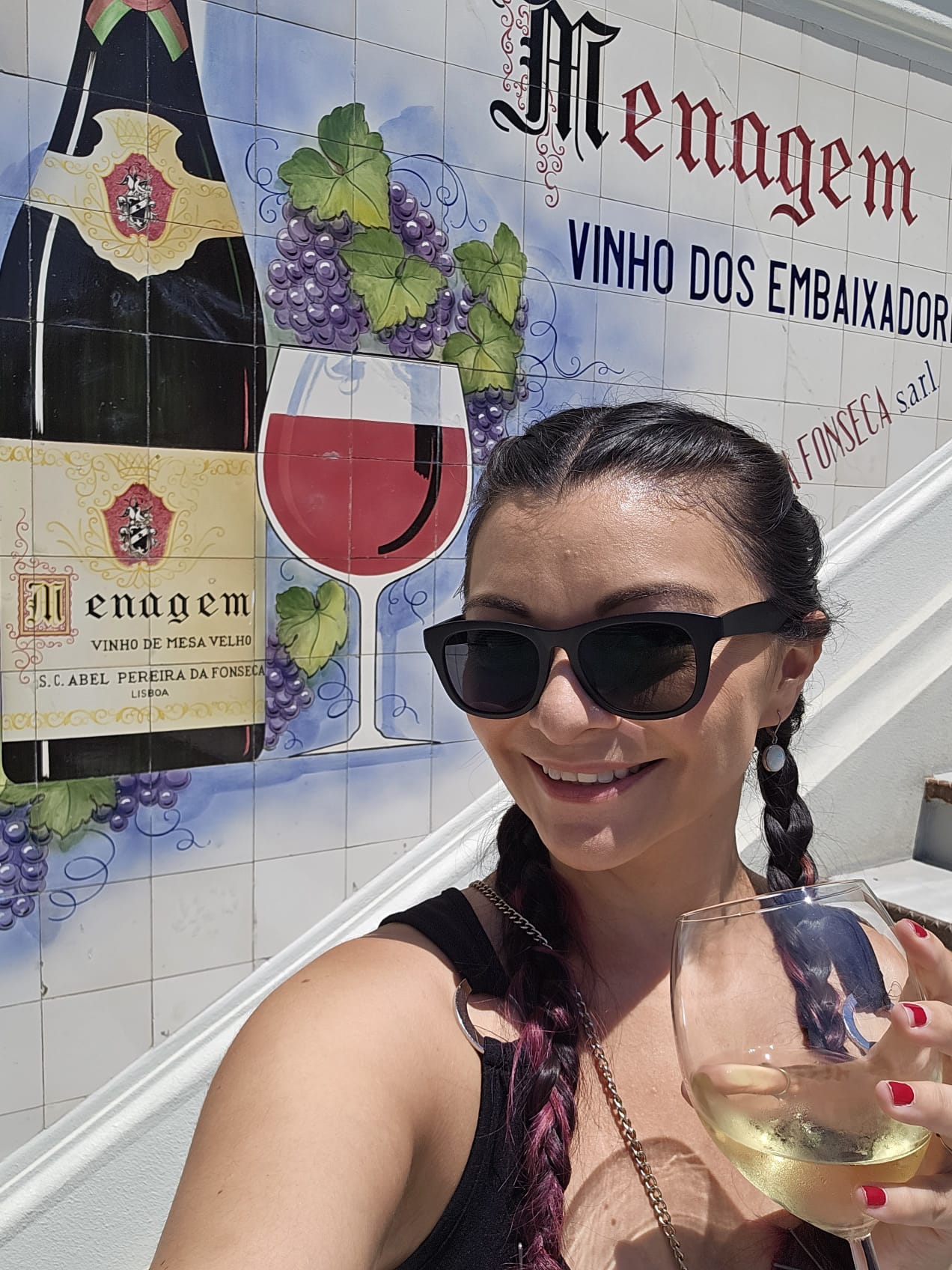 A person wearing sunglasses and braids smiles while holding a glass of white wine. They stand in front of a colorful mural featuring a wine bottle, grapes, and a glass of red wine, with text promoting "Menagem Vinho dos Embaixadores.".