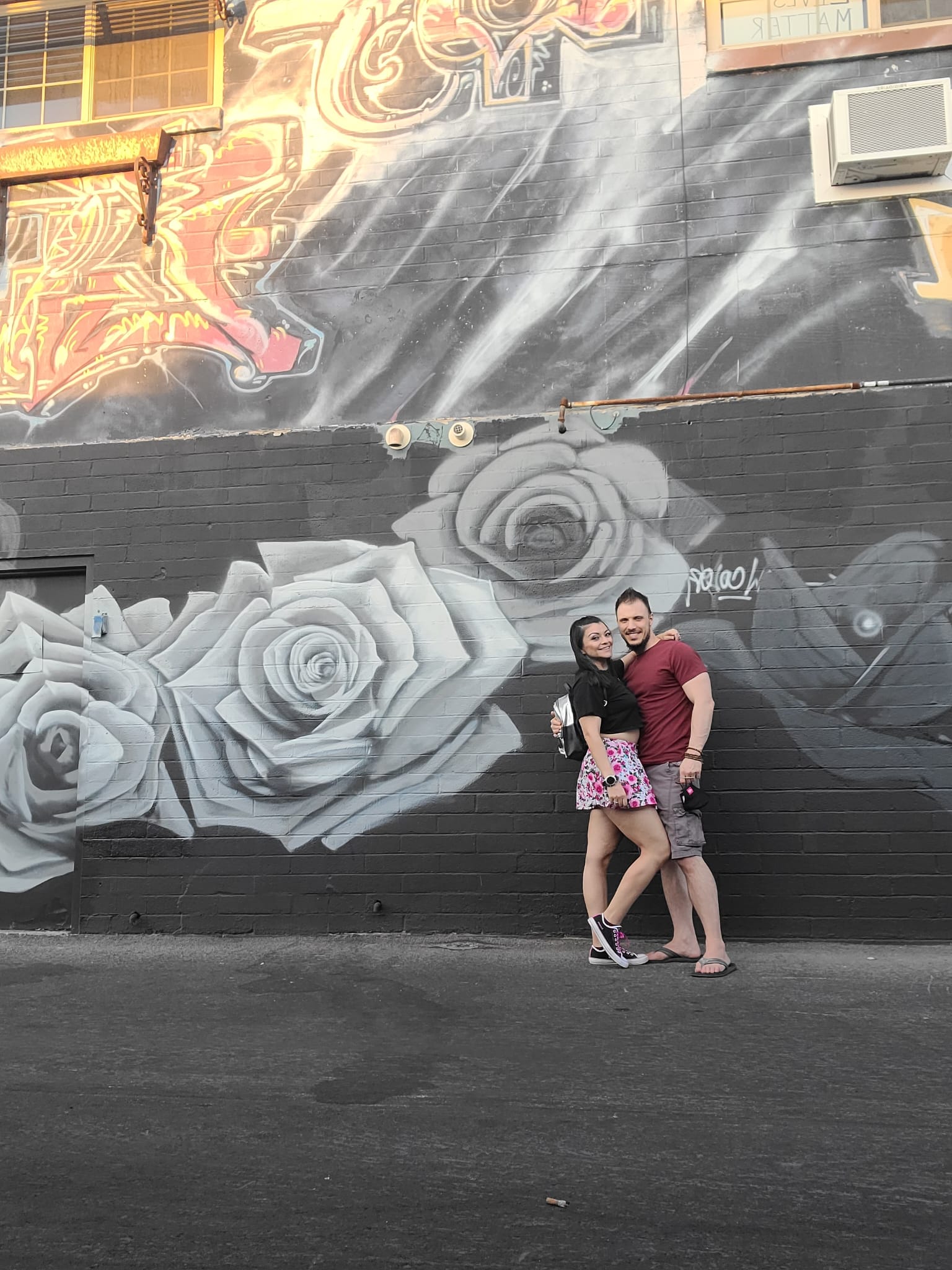 A couple poses happily in front of a vibrant mural featuring large, detailed roses and colorful graffiti. The woman wears a floral skirt and sneakers, while the man is in a maroon shirt and shorts. The mural's intricate design adds an artistic backdrop to the scene.