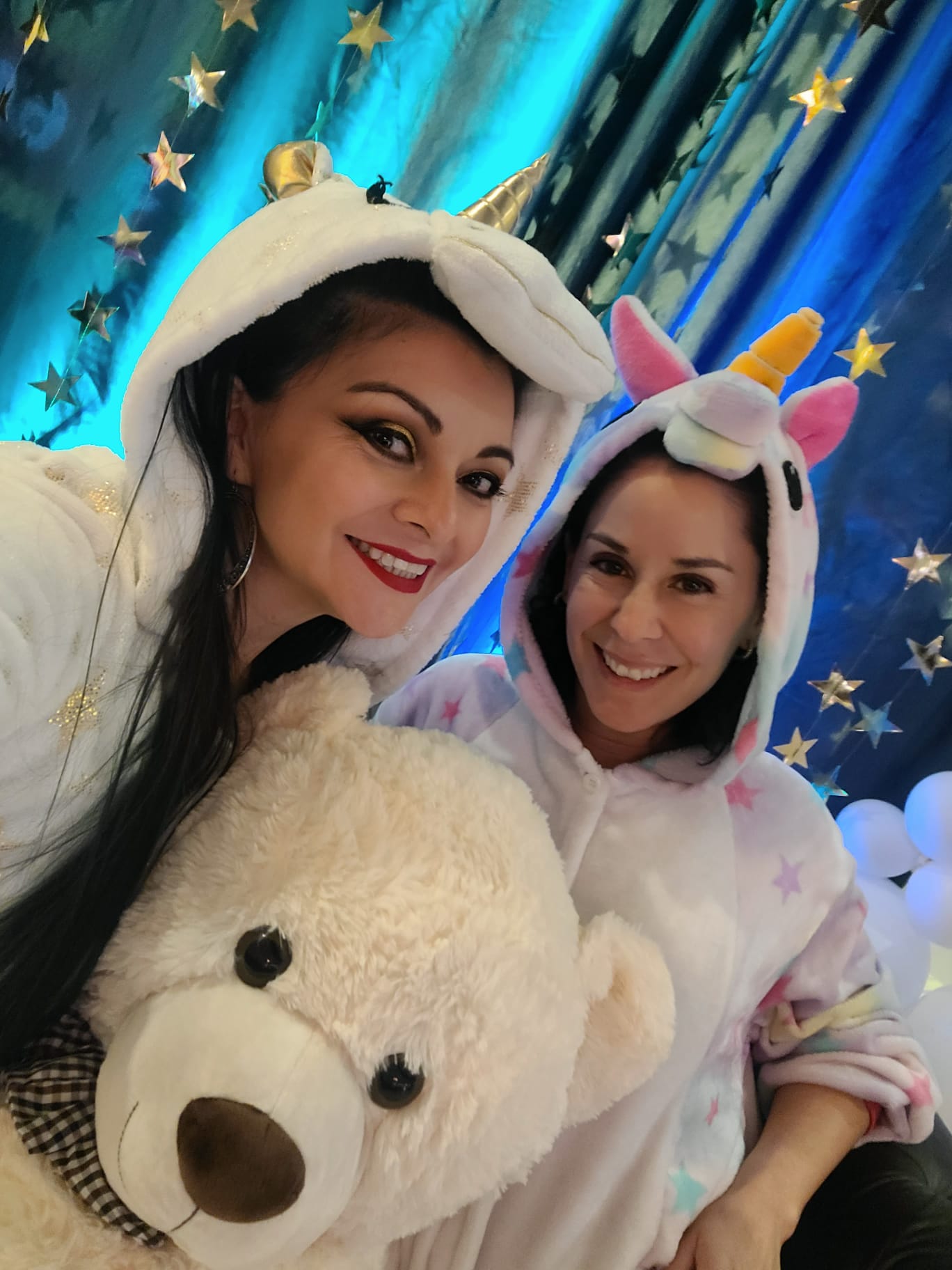 Two people in unicorn onesies smile brightly, holding a large plush teddy bear. The background features a blue curtain adorned with gold stars, creating a whimsical atmosphere.