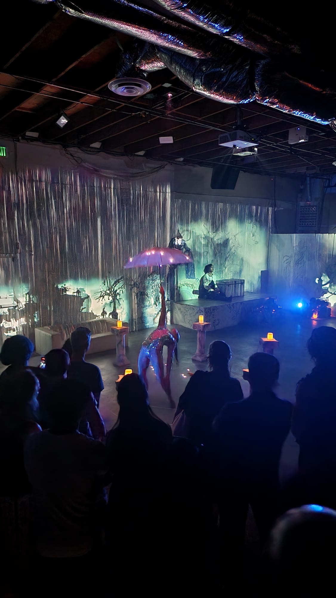A performer in a sparkling outfit dances gracefully under a glowing umbrella in a dimly lit room, surrounded by an audience. The setting features ambient lighting, decorative pillars with candles, and a backdrop of shimmering metallic curtains, creating a mystical atmosphere.