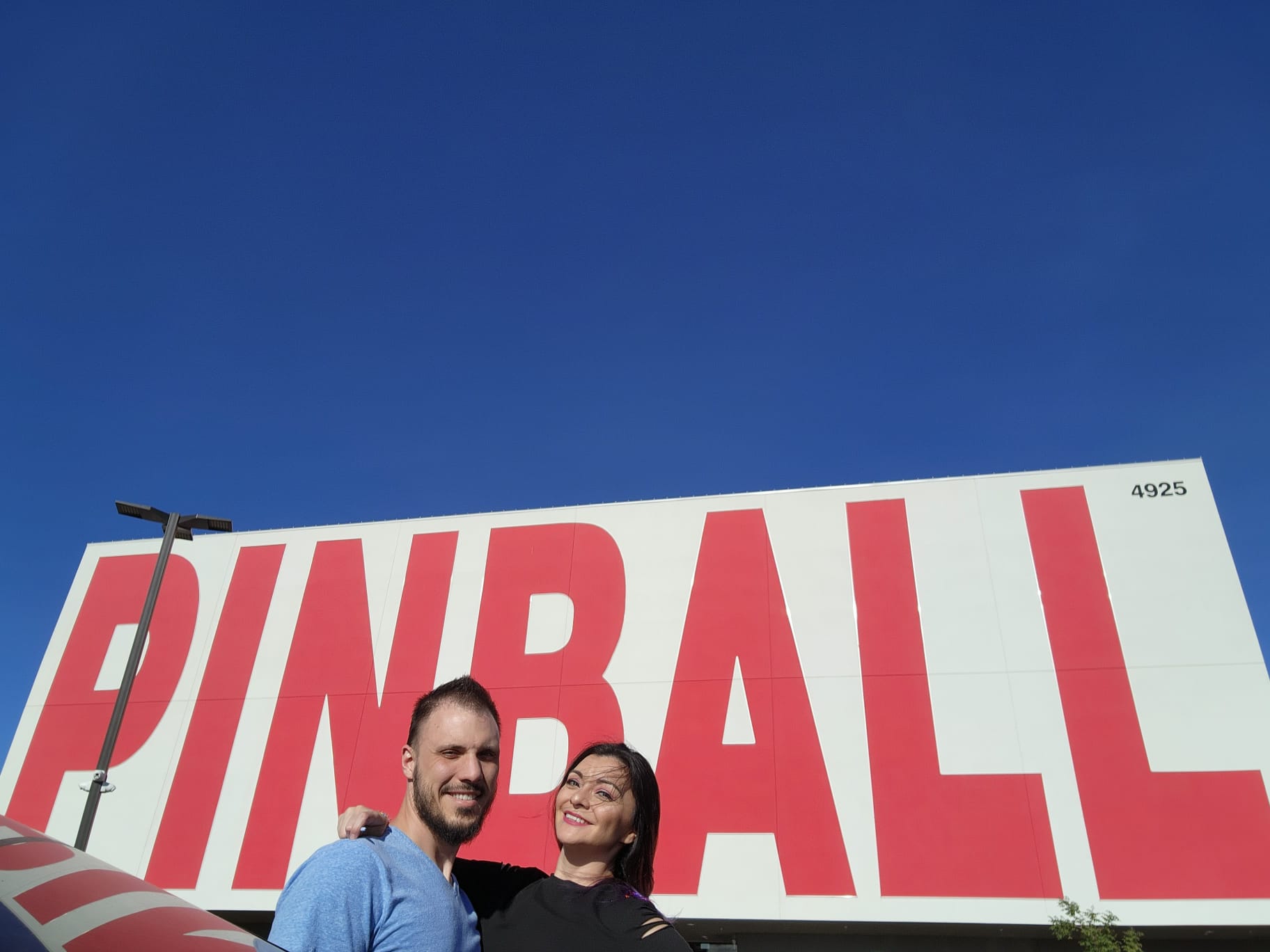A smiling couple stands in front of a large building with the word "PINBALL" in bold red letters on a white background. The sky is clear and blue, and a streetlight is visible nearby.