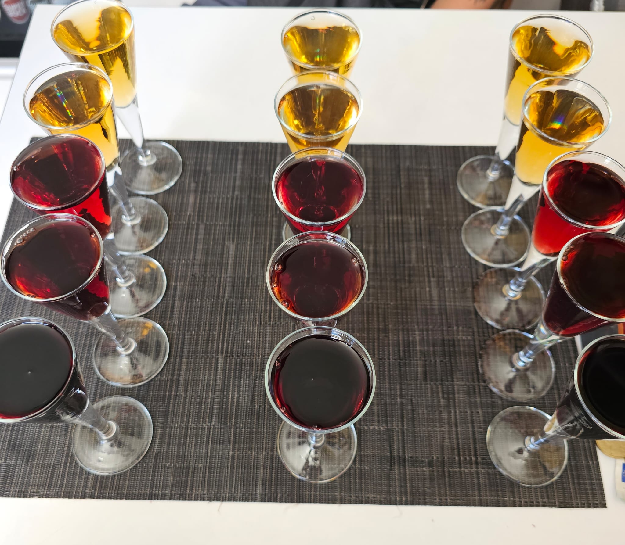 A neatly arranged set of champagne flutes filled with amber and deep red liquids sits on a textured dark placemat, creating a symmetrical and visually appealing pattern.