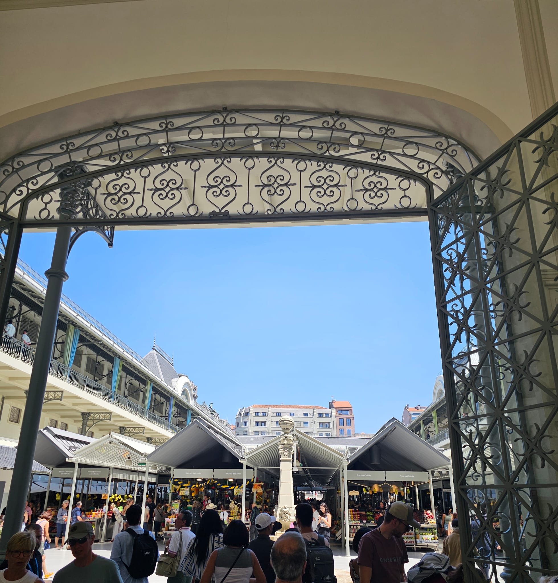 A bustling market scene viewed through an ornate, wrought-iron archway, with numerous people walking beneath the bright blue sky. The market stalls are covered with pointed roofs, and a stone statue stands prominently in the center. Surrounding buildings have classic architectural features.