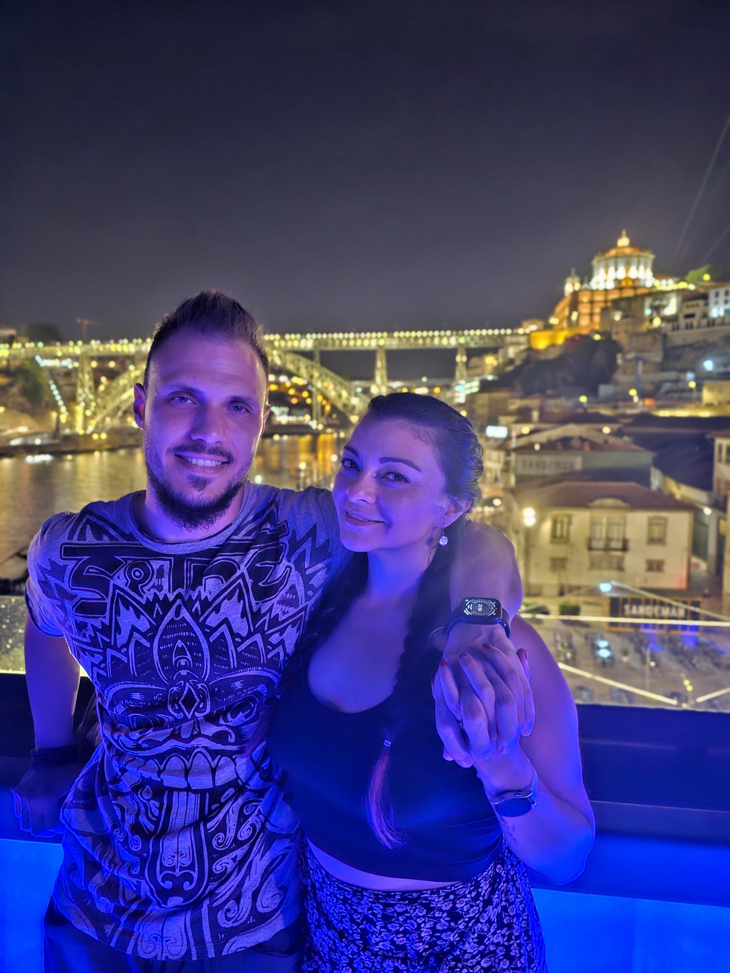 A smiling couple poses together against a nighttime cityscape, with a beautifully illuminated bridge and buildings in the background. The man wears a patterned shirt, and the woman, with braided hair, holds his hand while wearing a black top and patterned skirt.