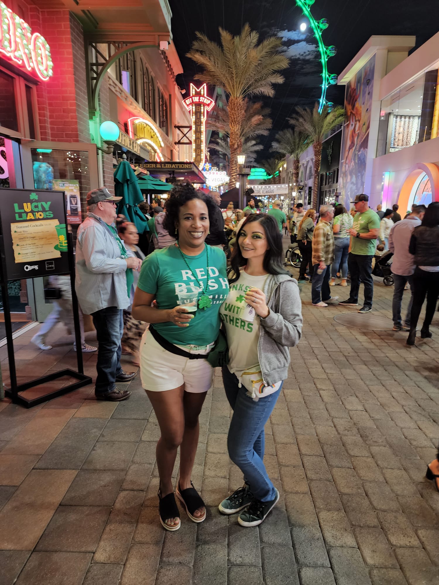 Two women stand smiling in a lively street scene, both holding drinks and wearing green shirts. The street is bustling with people, illuminated by neon signs and festive lights, with palm trees and a Ferris wheel visible in the background.