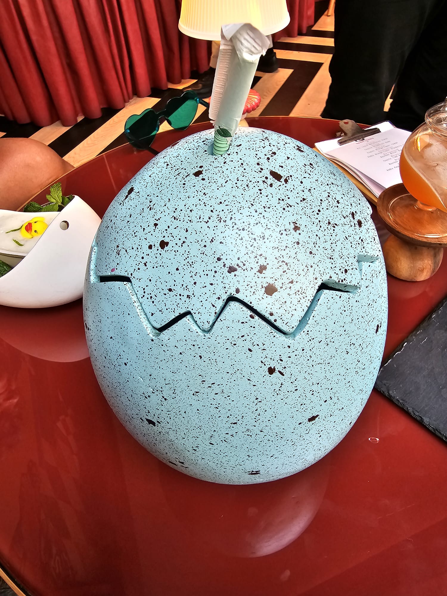 A speckled blue egg-shaped container with a zigzag opening sits on a red table, featuring a straw and napkin emerging from the top. Nearby, heart-shaped green sunglasses and a drink in a glass teapot add to the lively setting.