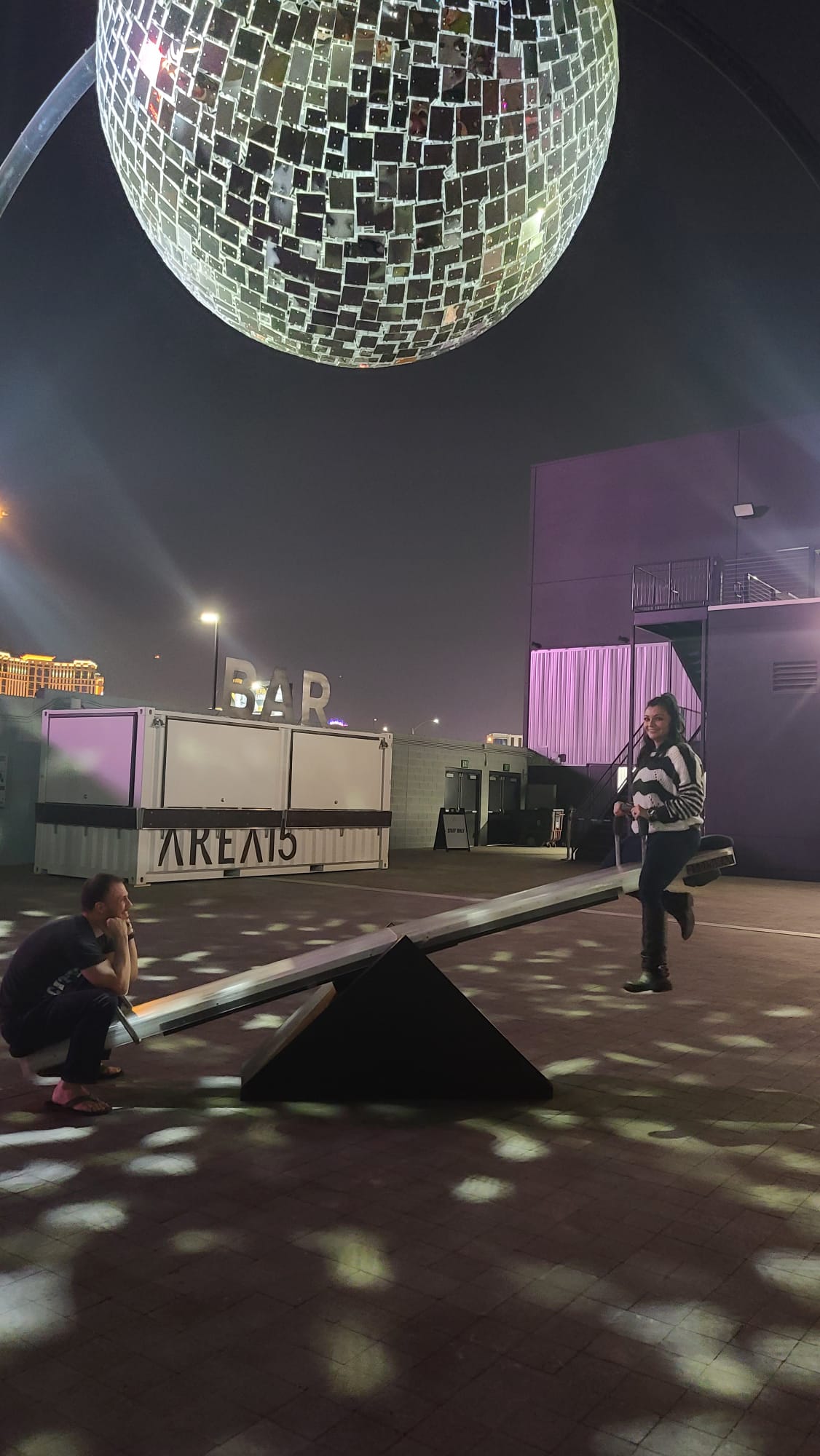 A person is sitting on one end of a seesaw while another person is standing on the opposite end, under a large reflective disco ball. The scene is illuminated with scattered light patterns, and in the background, a sign reads "BAR" next to a building marked "AREA15.".